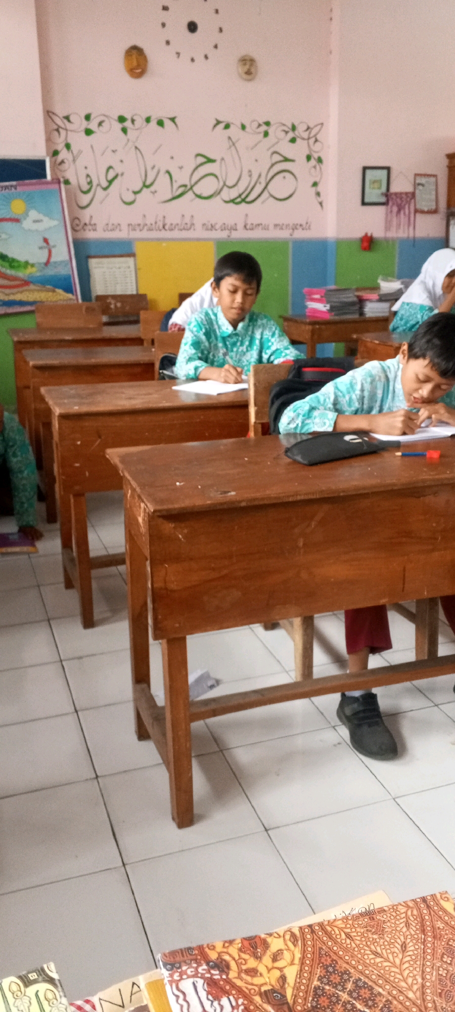 The image depicts a classroom scene with several young male students seated at wooden desks, appearing to be engaged in academic activities. The students are wearing similar patterned shirts, suggesting a school uniform. One student in the foreground is writing on a piece of paper, with a pencil and eraser on his desk. Another student behind him is also writing. The classroom has tiled flooring and a light-colored wall with decorative elements, including calligraphy and a clock. In the background, there are shelves with books and other materials. The overall environment appears to be that of a primary or secondary school in Lumajang, Indonesia.