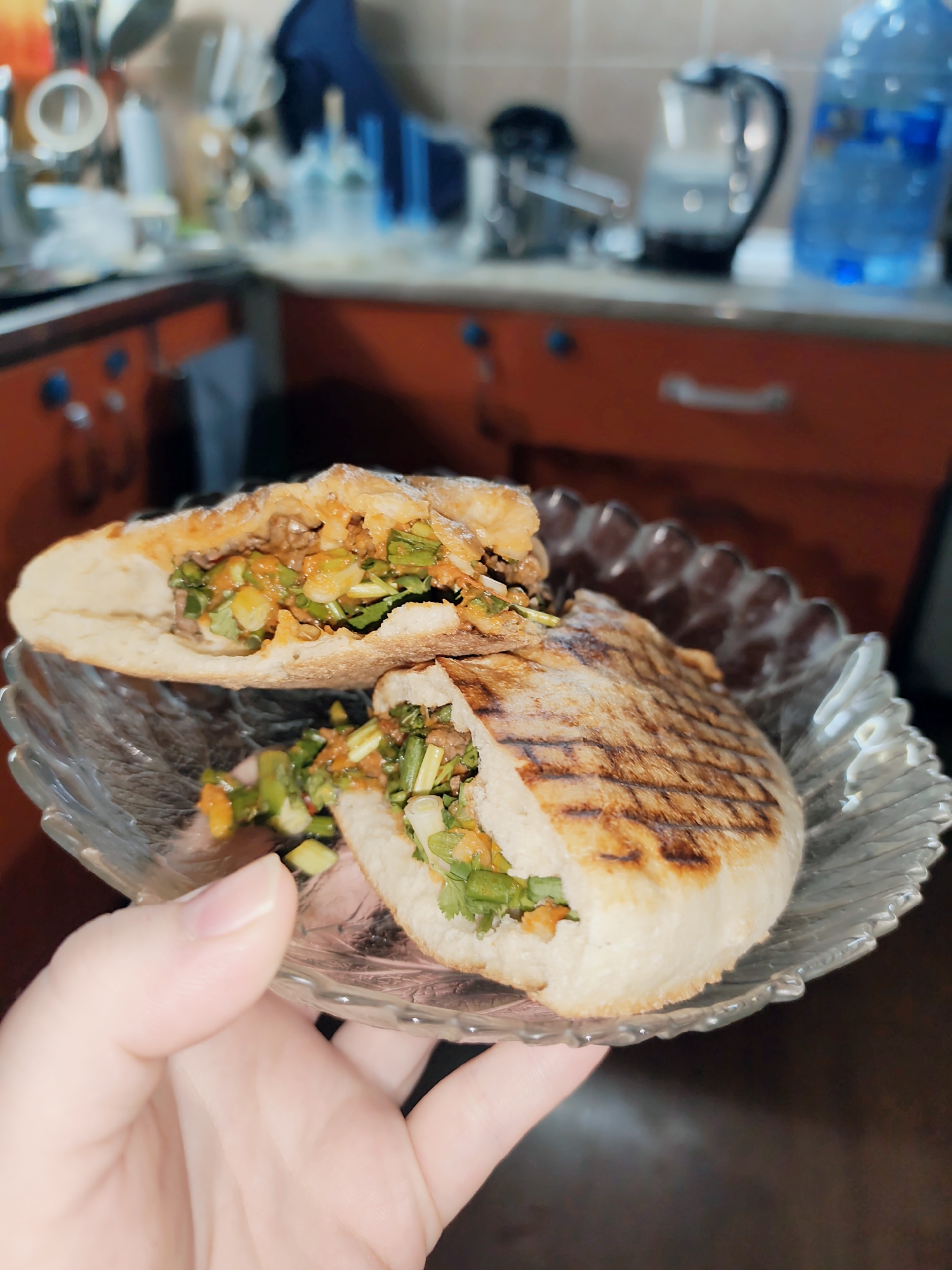 A close-up shot shows a person's hand holding a glass plate with a halved stuffed flatbread. The flatbread appears to be grilled, with visible grill marks on the exterior. The filling is a mixture of meat, green herbs, and possibly some vegetables. The background is blurred, showing kitchen elements like cabinets, bottles, and appliances. The scene is likely set in a kitchen in Fergana City, Uzbekistan.