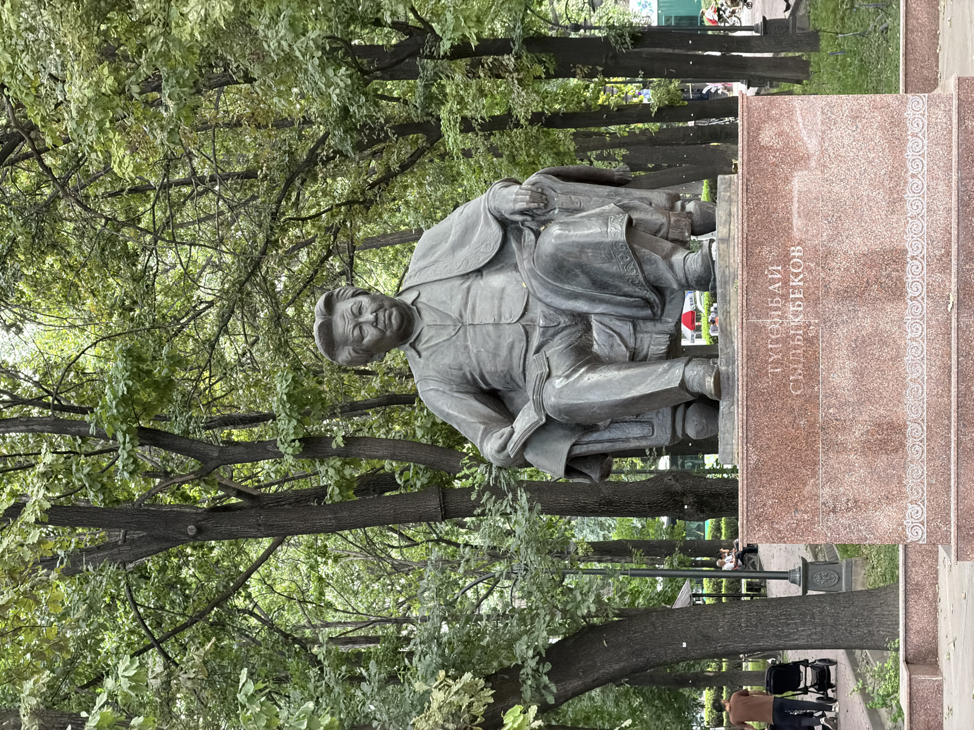 A large, weathered bronze statue of Togolok Moldo Sydykbekov, a Kyrgyz writer, is prominently displayed in Oak Park (Dubovy Park) in Bishkek, Kyrgyzstan. The male figure is depicted seated in an ornate armchair, wearing a collared shirt, vest, and a jacket draped over his shoulders, with his right hand holding an open book resting on his lap. The statue rests on a tall, rectangular pedestal constructed of reddish-brown speckled granite. The inscription "ТУГӨЛБАЙ СЫДЫКБЕКОВ" is carved in light-colored Cyrillic capital letters on the front face of the pedestal, above a horizontally oriented, repeating pattern of intricate white swirling motifs.
The statue is positioned within a dense arboreal environment, characterized by numerous mature trees with dark, textured trunks and abundant green foliage that extends throughout the background, indicating a park setting. Paved paths are visible around the base of the statue and extend into the background. On the left side of the image, a person in a brown shirt and dark pants pushes a black baby stroller on a paved path, moving away from the viewer. Further in the midground, several individuals are seated on a park bench beneath the trees. Ornate dark grey streetlights with decorative bases are interspersed among the trees. The scene is illuminated by natural daylight, suggesting an outdoor, daytime setting.