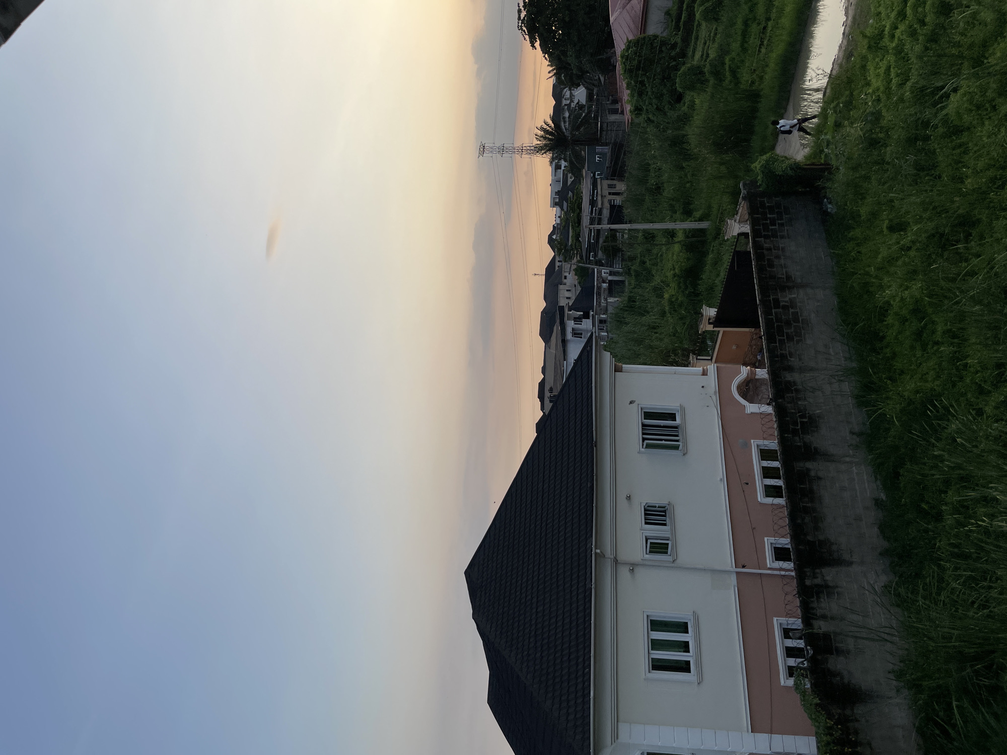 The image depicts a residential scene in Ado, Nigeria, during either sunrise or sunset. The sky exhibits a gradient of pale blue transitioning to a soft orange and yellow hue near the horizon. A few wispy clouds are scattered across the sky, with one elongated cloud notably present.
In the foreground, a large, modern house with a dark tiled roof and white and light pink exterior walls is prominently featured. The house has several rectangular windows with white frames. A low, brick wall topped with barbed wire runs along the front of the property, separating it from a patch of overgrown green grass and vegetation.
Beyond the immediate house, the cityscape reveals more residential buildings with similar architectural styles, interspersed with lush green foliage and palm trees. A tall, lattice-like electricity pylon stands in the mid-ground, with power lines stretching across the scene.
To the right of the image, a narrow, unpaved path runs alongside a puddle of water. A lone individual, appearing to be a young man wearing dark clothing and a backpack, is walking along this path away from the viewer. The overall environment suggests a semi-urban or suburban setting with a blend of developed properties and untamed natural growth.