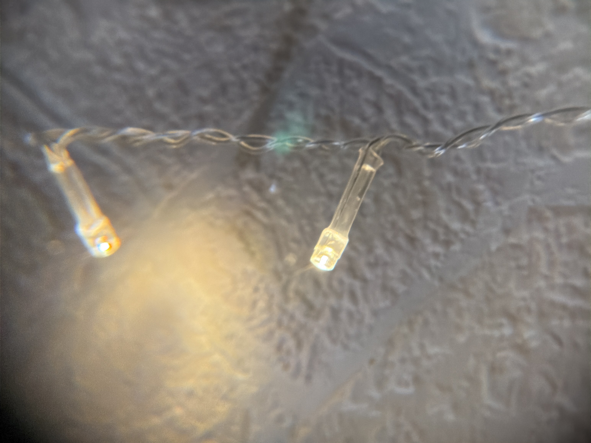 The image shows a close-up of two small, warm-white LED fairy lights connected by a thin, twisted wire. The background is a blurred, textured surface, possibly a wall or fabric, in shades of gray and off-white. There are no people or actions visible in the image. The scene appears to be indoors. The location cannot be determined based on the image content.