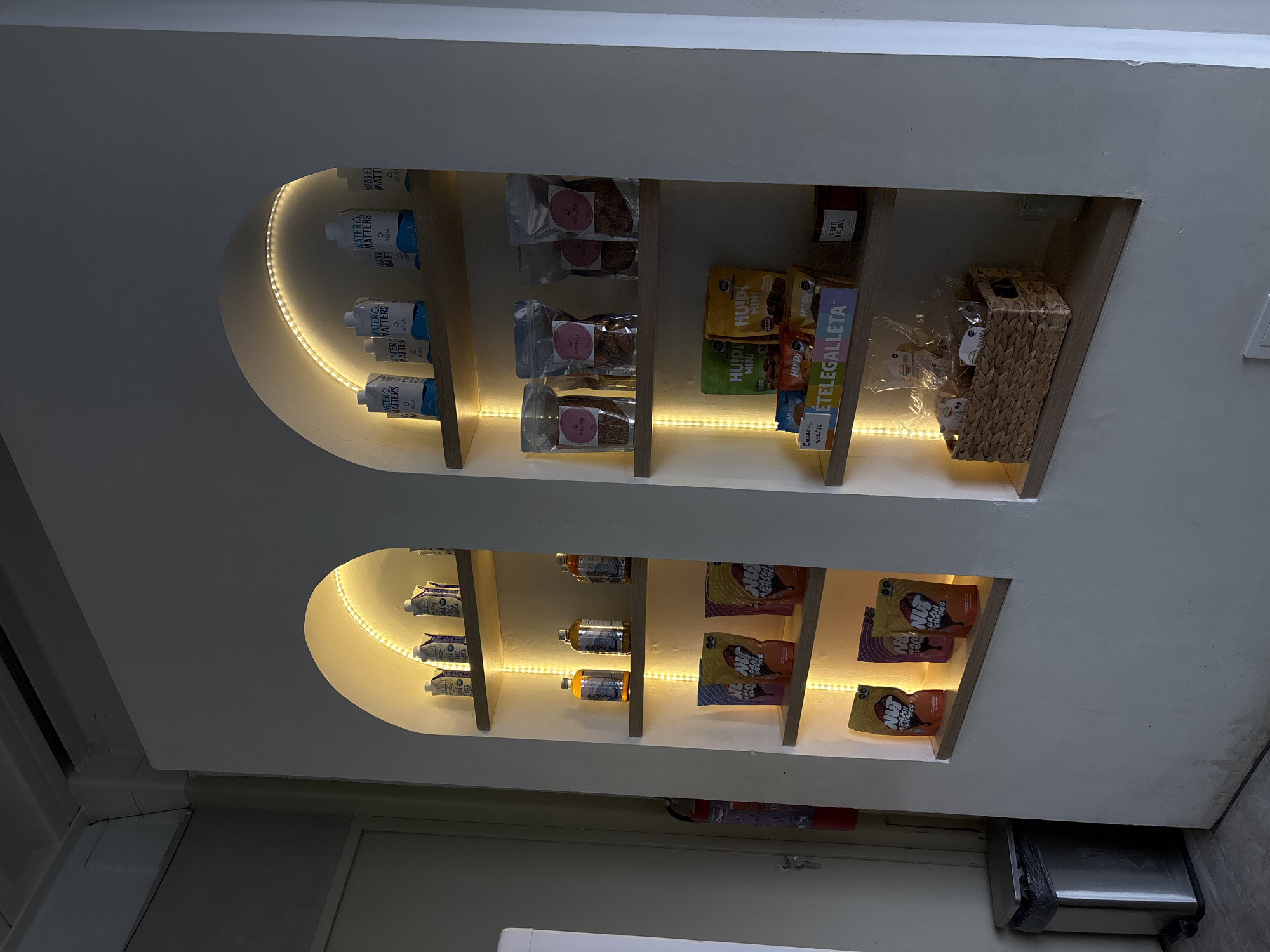 The image shows built-in shelves, possibly in a store, with various food and beverage items. The shelves are integrated into a white wall and have arched tops, each lined with LED strip lighting.
The upper shelf displays items such as "Water Matters" boxed water, packages labeled "Papalotl" (likely cookies or snacks), boxes of "Huipi Mini" and "Ételegalleta" cookies, a wicker basket filled with small packages, and a bottle of "Cider & Clove".
The lower shelf contains items like drink bottles, and packages of "Nut Damn Chance" snacks.
The image suggests the setting is a retail space, possibly a small store or cafe, located in San Bernardino Tlaxcalancingo, Mexico, based on the brand names and package designs. Other objects in the scene include an exit door, a fire extinguisher, and a trash can.