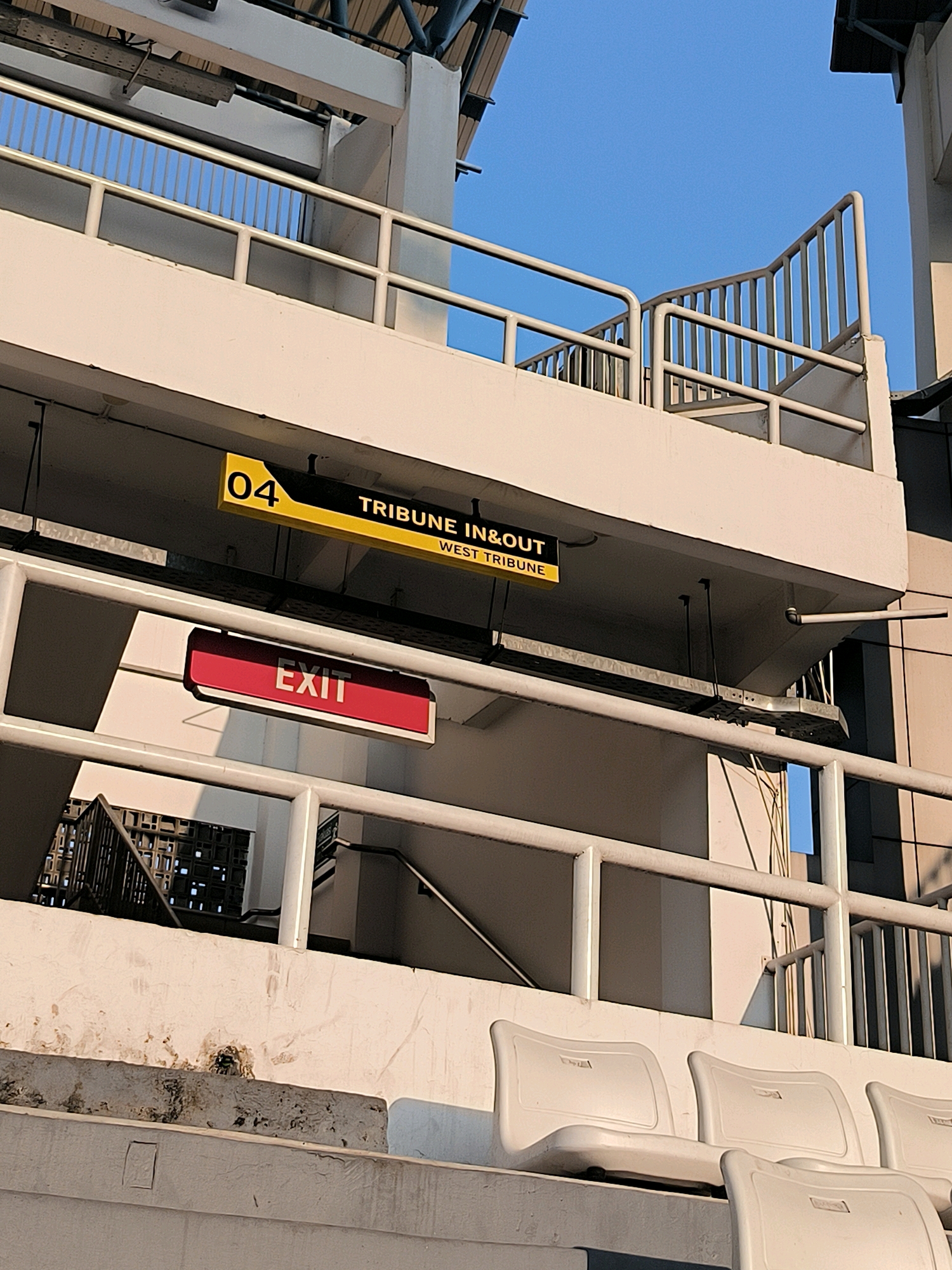 A low-angle, eye-level shot shows the exterior of a stadium structure under a clear blue sky. The prominent elements are white concrete and metal railings. A yellow and black sign reads "04 TRIBUNE IN&OUT WEST TRIUNE". Below this, a red and white illuminated sign displays the word "EXIT". A metal grate structure is visible on the left, partially obscured by a railing. In the foreground, empty white stadium seats are arranged in rows. The overall impression is of a functional, outdoor public space within an urban environment.
