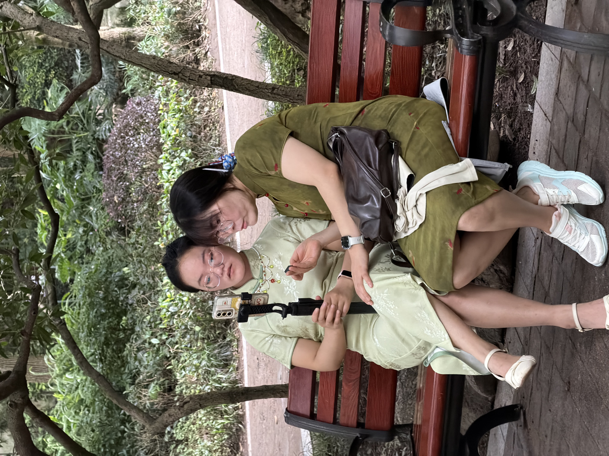 Two individuals are seated on a red park bench in a shaded outdoor area, likely a park, in Jiulongpo District, China. The person on the left, wearing a light green traditional Chinese dress and white flat shoes, is holding a smartphone mounted on a black selfie stick. The person on the right, in a darker green traditional Chinese dress, is wearing white sneakers and has a brown handbag resting on their lap. Both individuals are looking at the phone. The background consists of lush green foliage and trees, suggesting a natural, park-like setting. The bench itself is made of wood with dark metal legs and armrests.