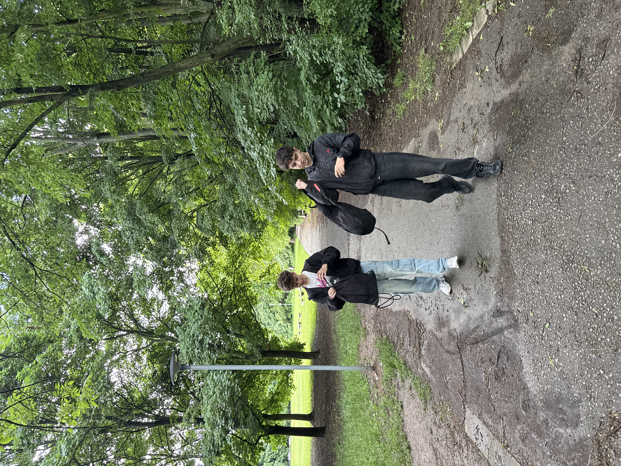 The image captures two young men standing on a winding dirt and gravel path within a verdant park or wooded area in Sofia, Bulgaria. The path shows patches of dampness or recent moisture, suggesting it might have rained recently. Lush green trees and dense bushes flank both sides of the path, creating a natural canopy that allows bright daytime light to filter through.
The man in the foreground, with lighter, curly hair, is dressed in a black jacket over a white t-shirt, light-wash jeans, and white sneakers. He is holding a black bag and looking down, seemingly engaged in retrieving or adjusting something within it.
Slightly behind him, the second man, with dark hair, wears a black jacket, dark pants, and black athletic shoes. He is also holding a black bag and looking downwards, mirroring the action of the first individual.
A grey metal pole, likely a lamppost, stands between the two men, positioned beside the path. Both individuals appear to be casually paused or walking, focused on their bags. The scene conveys a calm, outdoor moment in what appears to be a well-maintained urban green space. No visible text is present in the image.