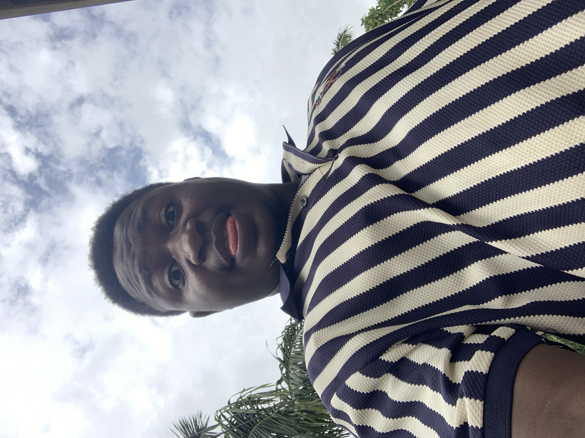 A low-angle shot shows a person, likely male, with dark skin, short dark hair, and wearing a striped navy and white polo shirt. The person is looking down at the camera, and their tongue is slightly visible. The background consists of a cloudy sky with patches of blue and some green palm leaves partially visible. The location is purportedly Akwanga, Nigeria, but there are no visual cues in the image to confirm the location.