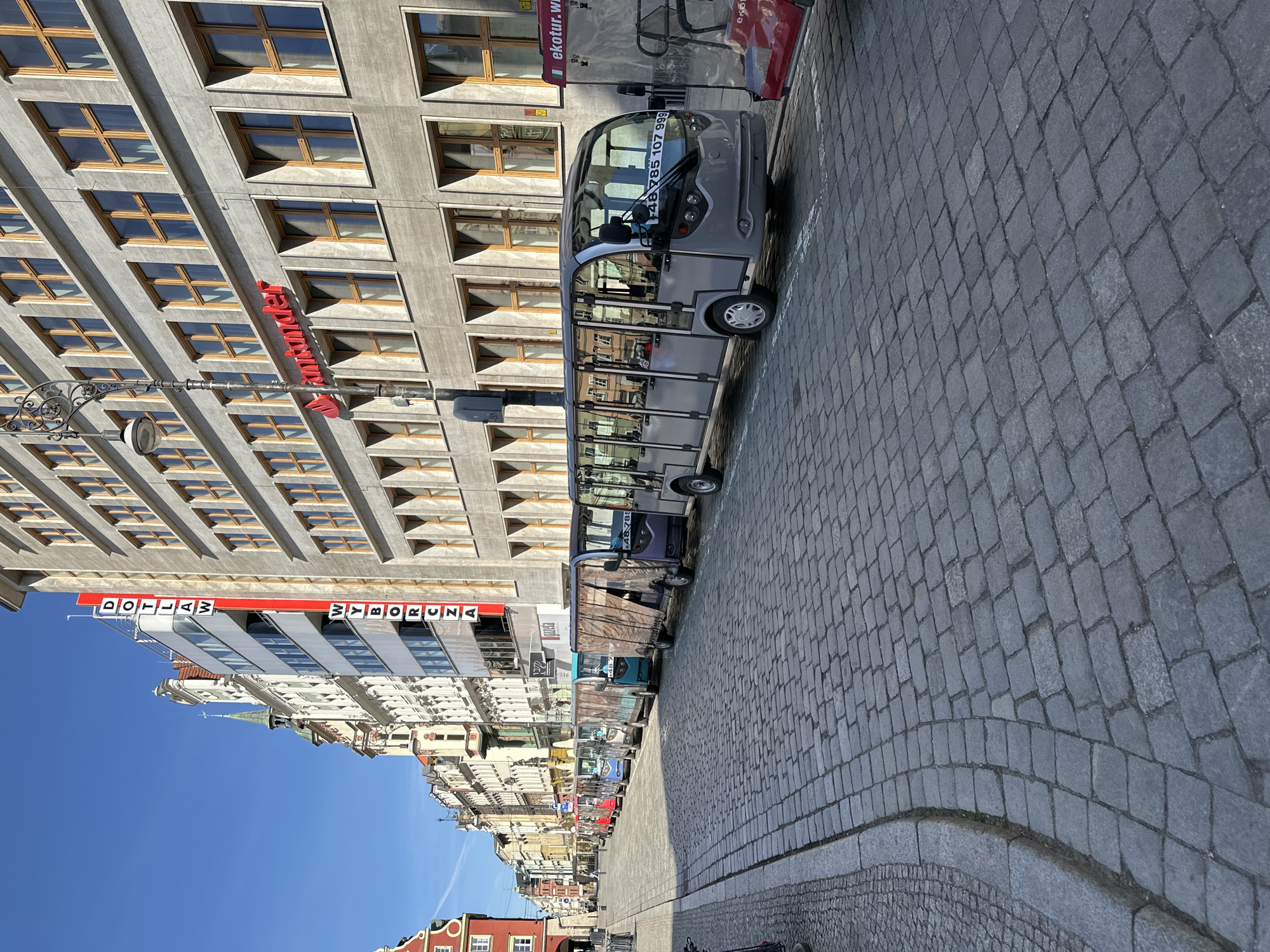 The image depicts a street scene in Wrocław, Poland. A line of small, electric tourist vehicles is parked along a cobblestone street. The vehicles appear to be designed for sightseeing tours. A tall, modern building with a Santander sign is visible on the left. Other buildings line the street, suggesting an urban environment. The sky is clear and blue. There are no people prominently visible in the frame. The overall scene captures a typical street with tourist-oriented transportation in a European city.