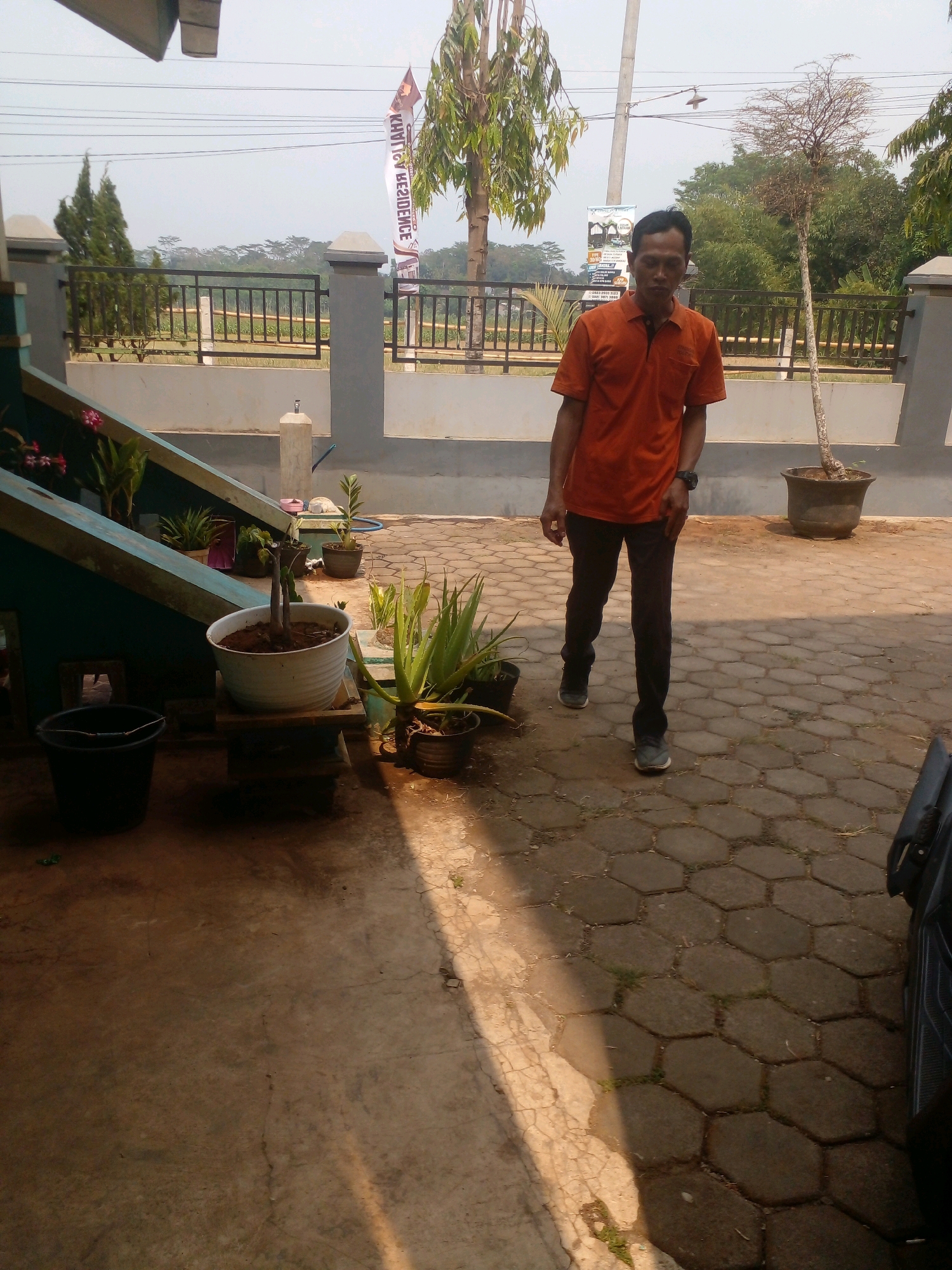 A man is standing outside in front of a building. He is wearing an orange short-sleeved collared shirt and dark pants. He has dark hair and is looking towards the camera. In the background, there is a fence with plants behind it, and trees and sky are visible. To the left of the man, there are several potted plants. The ground is paved with hexagonal stones and concrete. A black bucket is on the left side of the image. A flag with text on it is visible in the background. The location appears to be in the city of Banyuputih, Indonesia.