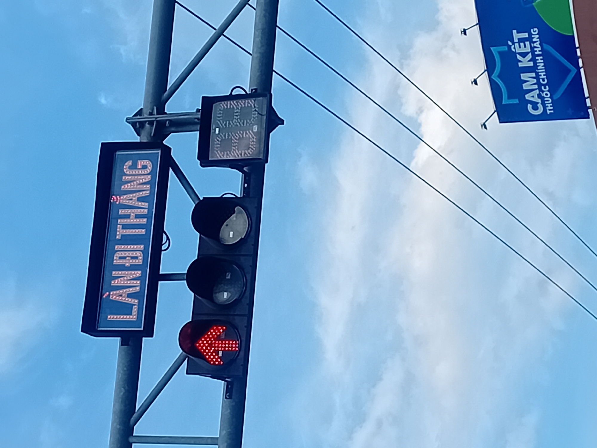 The image shows a traffic signal in Da Nang, Vietnam, displaying a red arrow pointing upwards, indicating that traffic should proceed straight. Above the signal, a blue sign with illuminated white letters reads "LÀN ĐI THẲNG," which translates to "STRAIGHT LANE." To the right of the traffic signal, there is a digital display showing the number "68" in a segmented font. In the background, power lines are visible against a cloudy blue sky. Below the traffic signal, a blue billboard with white text states "CAM KẾT THUỐC CHÍNH HÃNG," meaning "GUARANTEED GENUINE MEDICINE." The scene depicts a typical urban traffic management setup.