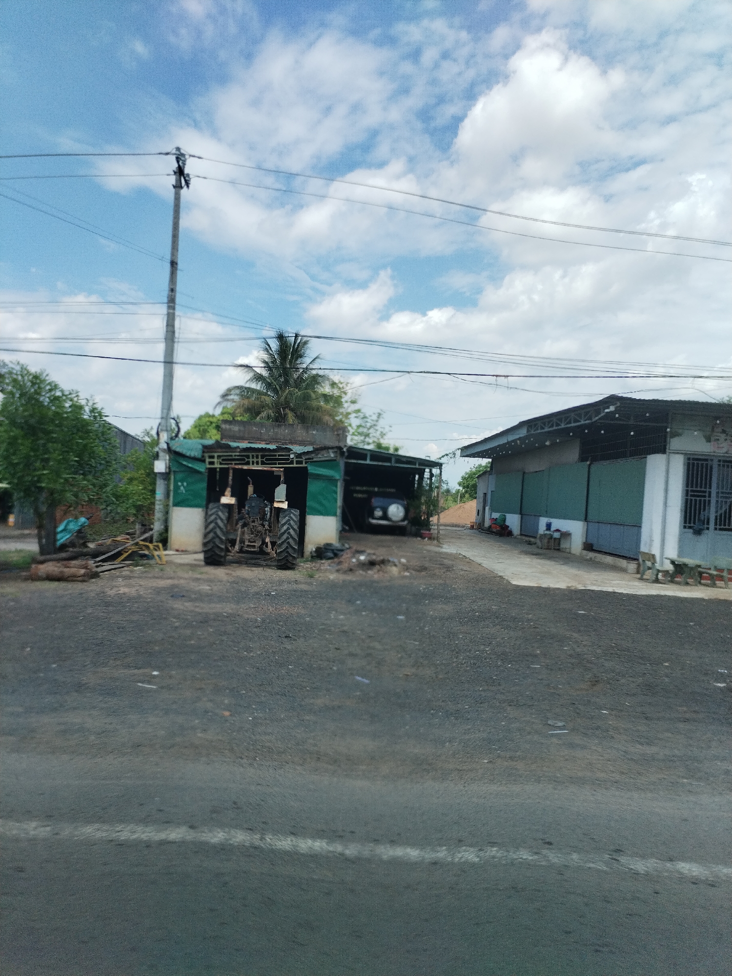 The image depicts a rural scene in Ia Le, Vietnam. A weathered tractor is parked under a dilapidated open shed. A utility pole with visible electrical wiring stands nearby. To the right, a long, low building with green paneling and a corrugated roof can be seen. The foreground consists of an unpaved area, likely a road, with a faded white line marking the edge. The sky is partly cloudy.