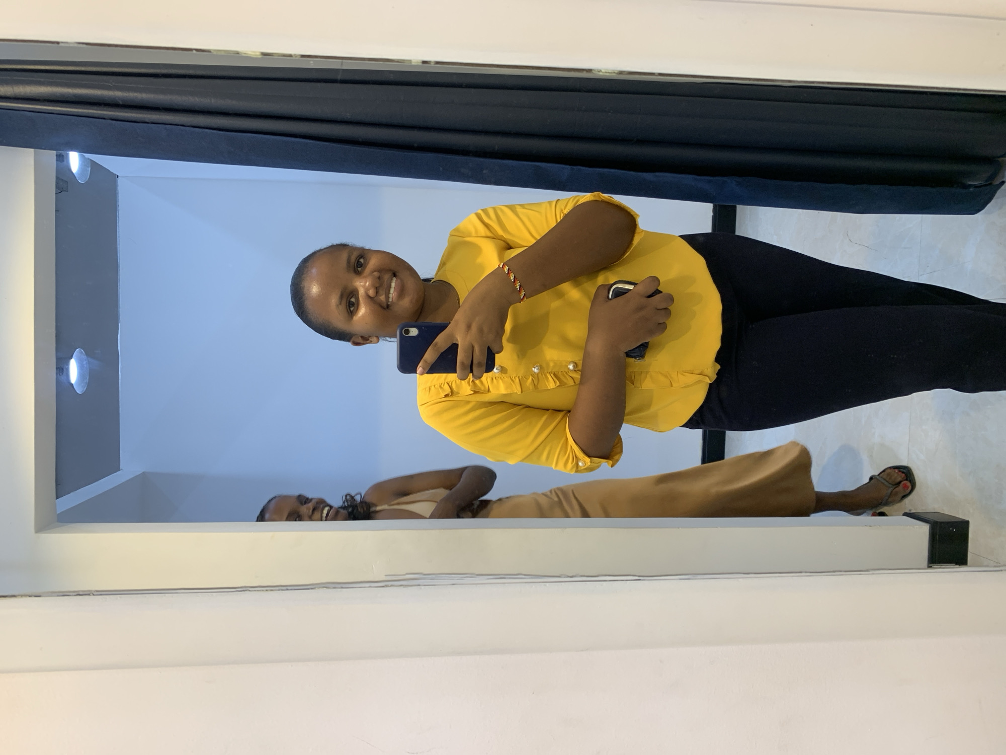 A woman in a yellow blouse and black pants takes a selfie in a mirror. In the mirror's reflection, another woman is visible wearing a tan dress. The location appears to be indoors, possibly a shop or dressing room, with a blue curtain hanging in the background. The exact location in Dar es-Salaam cannot be determined from the image.