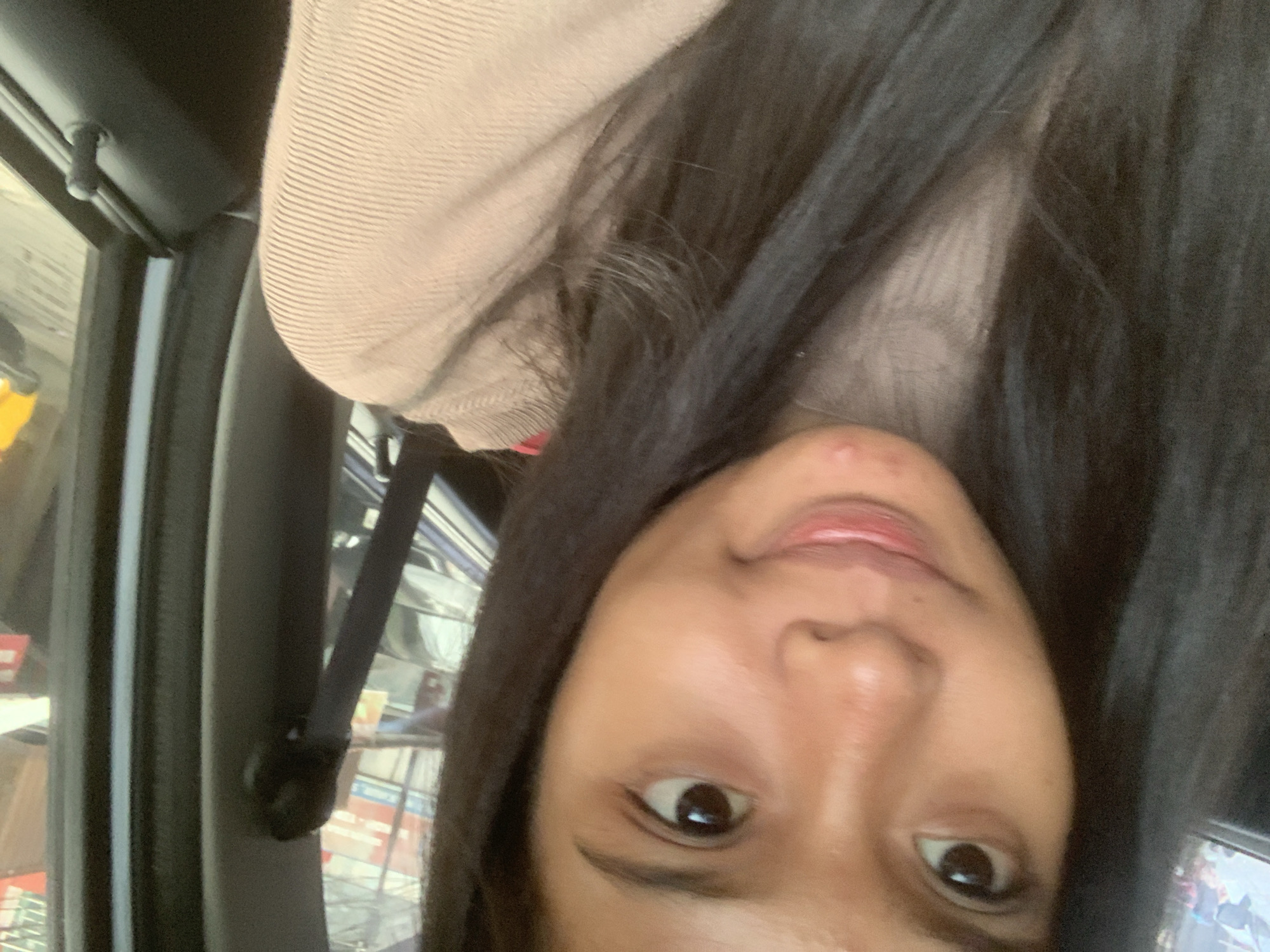 A person is sitting inside a vehicle. The individual has dark hair, brown eyes, and light brown skin. They are wearing a beige, textured top. The interior of the vehicle includes a gray door frame, a seatbelt, and a glimpse of a blurry background. The photograph does not provide enough information to place the location in the city of Samarinda, Indonesia.