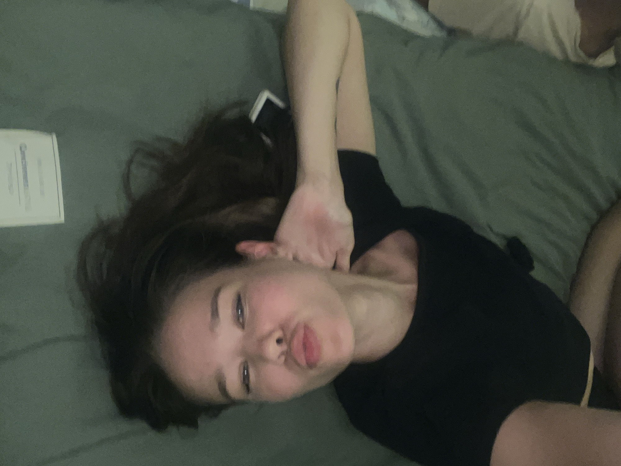 A woman is lying on a green bed. She has dark hair and is wearing a black shirt. She is making a kissing face and has one hand raised near her face. A white object is visible in the upper left of the frame. The location is not discernible based on the image.