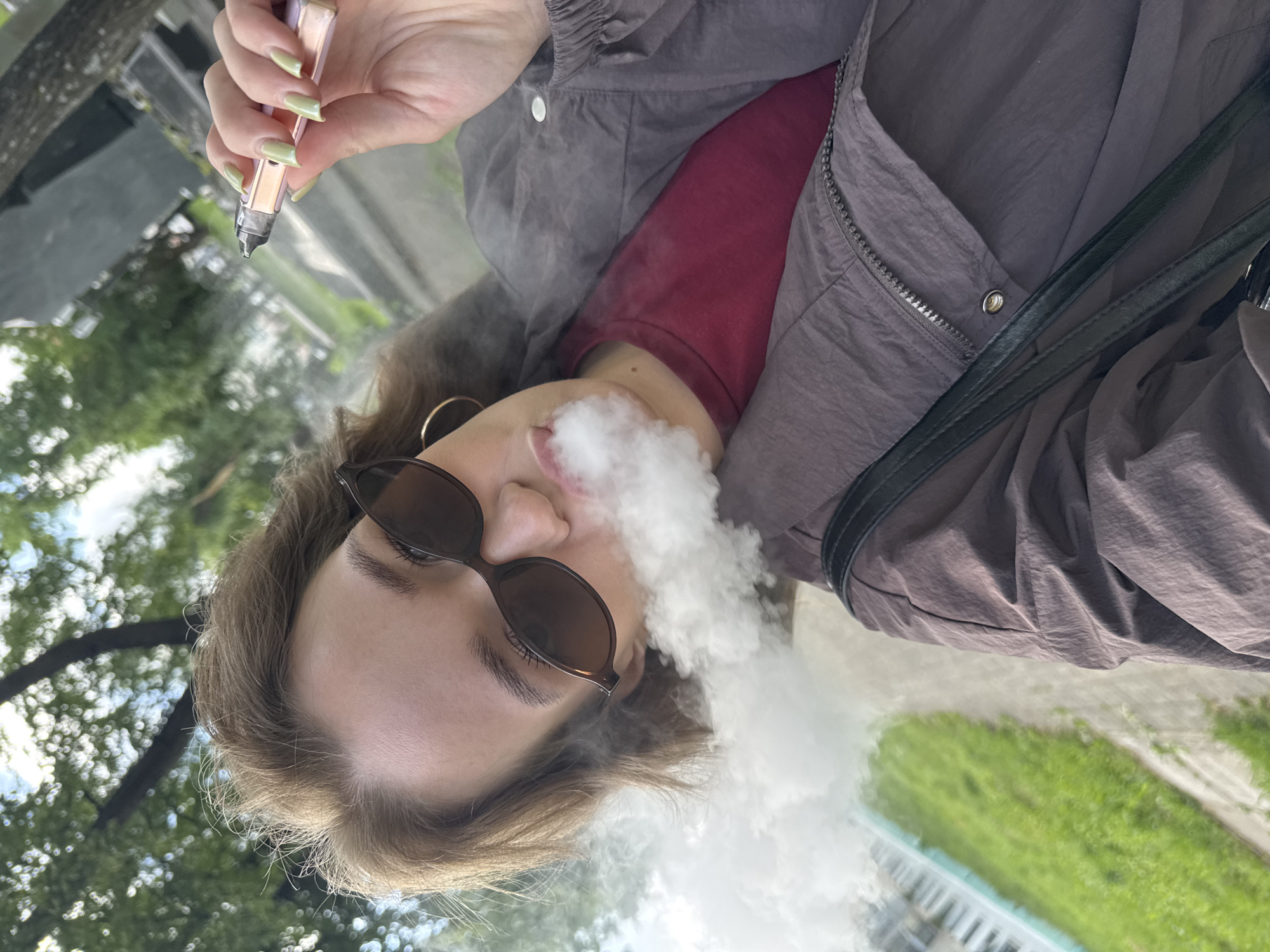 A young woman with light brown hair and sunglasses is exhaling a cloud of vapor in an outdoor, urban setting. She is holding a rose gold vape pen in her right hand, which is near her face. She is wearing a dark maroon t-shirt and a dark brown jacket. The background suggests a park or a street with trees and a path. The lighting indicates it is daytime.