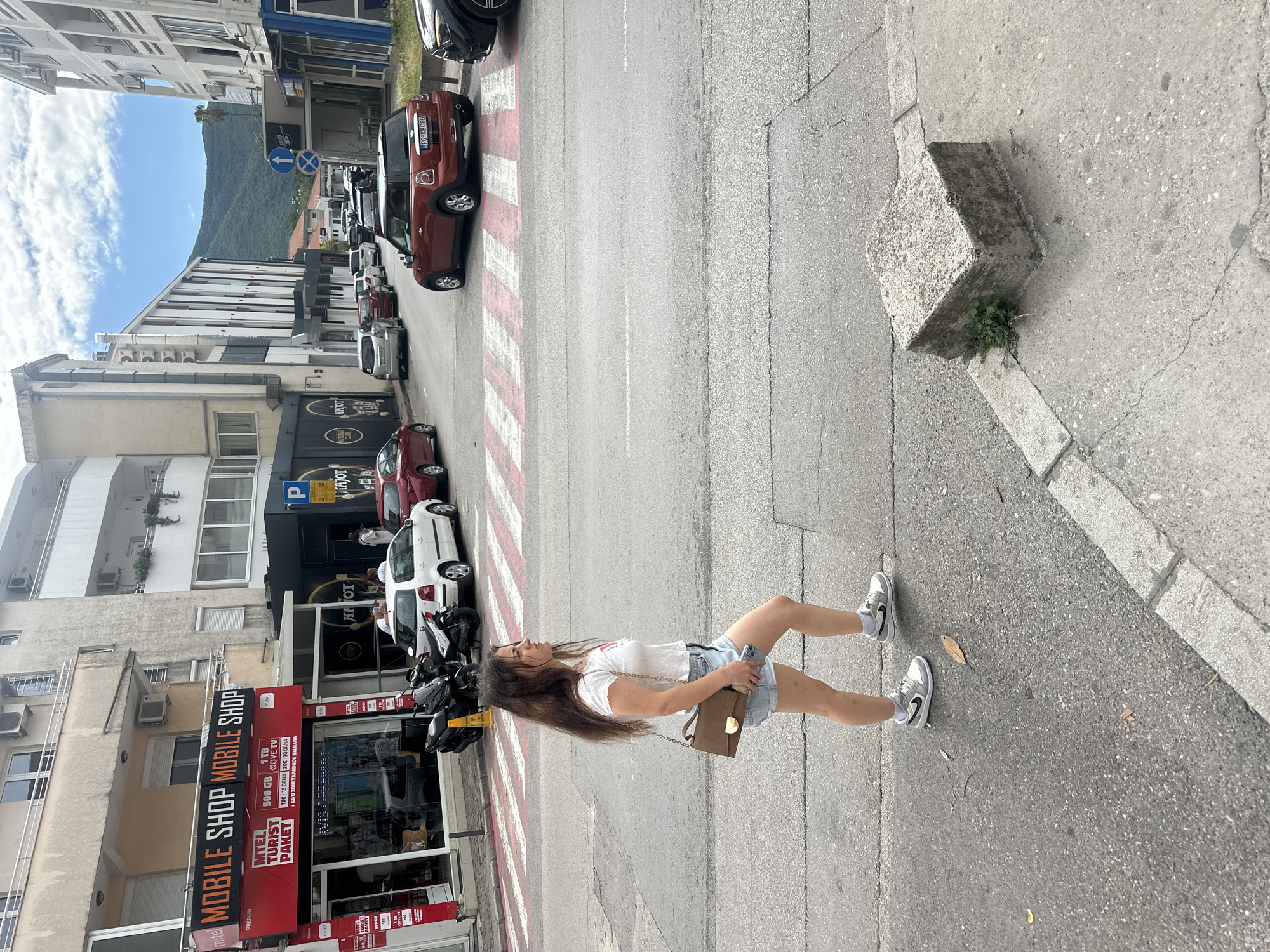 The image captures an urban street scene in Budva, Montenegro, on a bright, sunny day. The central subject is a young woman with long brown hair, wearing a white t-shirt, denim shorts, and white sneakers. She is holding a smartphone in her left hand and a brown shoulder bag, and appears to be walking along a sidewalk or near a street, looking slightly upwards and to her left.
The street features a red and white striped pedestrian crossing and a continuous white line, with several cars parked along its edge and others visible further down the road. A reddish-brown car with the license plate "PG K8086" is prominently parked next to the crossing. On the opposite side, two motorcycles or scooters are parked near a commercial building.
The surrounding environment consists of multi-story buildings, some appearing residential with balconies and air conditioning units, and others housing ground-level businesses. A "MOBILE SHOP MOBILE SHOP" with a red sign is visible, displaying text like "MTEL TURIST PAKET," "PREPAID," "500 GB 1 TB," "MOVE TV," and pricing information such as "15€ 15 DANA 20€ 30 DANA + GB U ZONI ZAPADNOG BALKANA." Another business sign for "KAJOT CLUB" and "AUTOMAT KAJOT" is also present, along with a blue "P" parking sign.
In the background, lush green mountains rise under a blue sky dotted with white clouds, suggesting a scenic location. A rough concrete block sits on the corner of the sidewalk, with some small green plants growing beside it. The overall impression is a bustling daytime street in a Mediterranean town.
