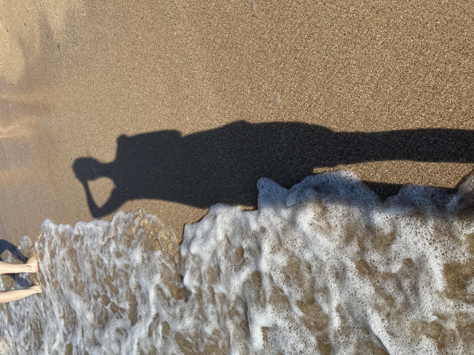 The image captures a perspective from the shore of Golubitskaya, Russia, showing a sandy beach with the gentle lapping of shallow waves. In the foreground, a shadow of a person with their arm raised to their head is cast on the damp sand. To the left, the foamy edge of the sea washes over the shore, and the feet of one or two people are visible standing in the shallow water. The sand is a light brown color, speckled with small dark grains, and reflects the warm sunlight. The overall scene depicts a peaceful moment at the seaside.