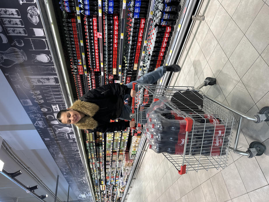 An adult female is depicted in a brightly lit supermarket aisle, identified as being in Sochi, Russia. She is actively balancing on the rear edge of a metal shopping cart, holding its red plastic handle with both hands, with her feet lifted from the light-tiled floor. Her expression is a broad smile. She wears a black bomber jacket with a large, light brown fur collar and blue jeans. The shopping cart contains at least three multi-packs of "Добрый cola" (Dobry cola) in dark bottles, sealed in clear plastic. Supermarket shelves extend along both sides of the aisle. The right-side shelves are heavily stocked with rows of red-labeled "Добрый cola" and blue-labeled bottled beverages. The left-side shelves display various other packaged goods. A dark wall mural with white line-art illustrations of beverages and barware is visible above the shelves. Overhead lighting fixtures are present.