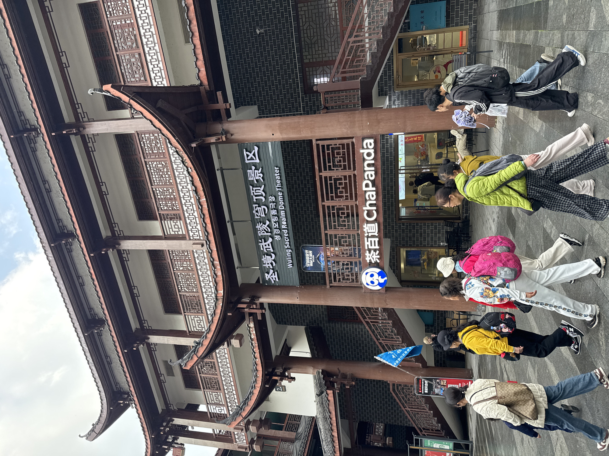 The image depicts a street scene in Wulingyuan, China, focusing on a multi-story building featuring traditional Chinese architecture. The building exhibits dark-tiled, multi-tiered curved roofs, exposed wooden beams, and white upper-level walls with ornate wooden lattice railings and windows. The ground floor is constructed with dark grey brick. A prominent sign on the facade reads "圣境武陵穹顶景区" and "Wuling Sacred Realm Dome Theater" in English, with "ChaPanda" (茶百道) branding below it, indicating a retail establishment. An external staircase with dark risers and wooden handrails ascends on the right side of the building.
On the paved sidewalk in the foreground, approximately ten individuals are visible. From left to right:
* An adult female in a beige quilted jacket, blue jeans, and a light-colored shoulder bag walks alongside a small child in dark clothing.
* An adult female in a bright yellow top, black pants, and a black backpack walks ahead.
* Two adult females are positioned next to each other, both carrying bright pink backpacks. One wears a white and multi-colored tracksuit; the other wears a light-colored top and beige pants.
* An adult female in a bright green quilted jacket, plaid pants, and a black backpack walks to the right.
* Another adult female in a mustard-yellow top and light-colored wide-leg pants walks behind the woman in green.
* A young adult male in a black Adidas tracksuit and a grey backpack holds a light blue item, possibly an umbrella or garment.
A blue flag on a pole is visible on the left side of the frame, near the building's entrance. Outdoor seating is partially visible outside the ChaPanda store. The sky is overcast, and the ground appears slightly damp.