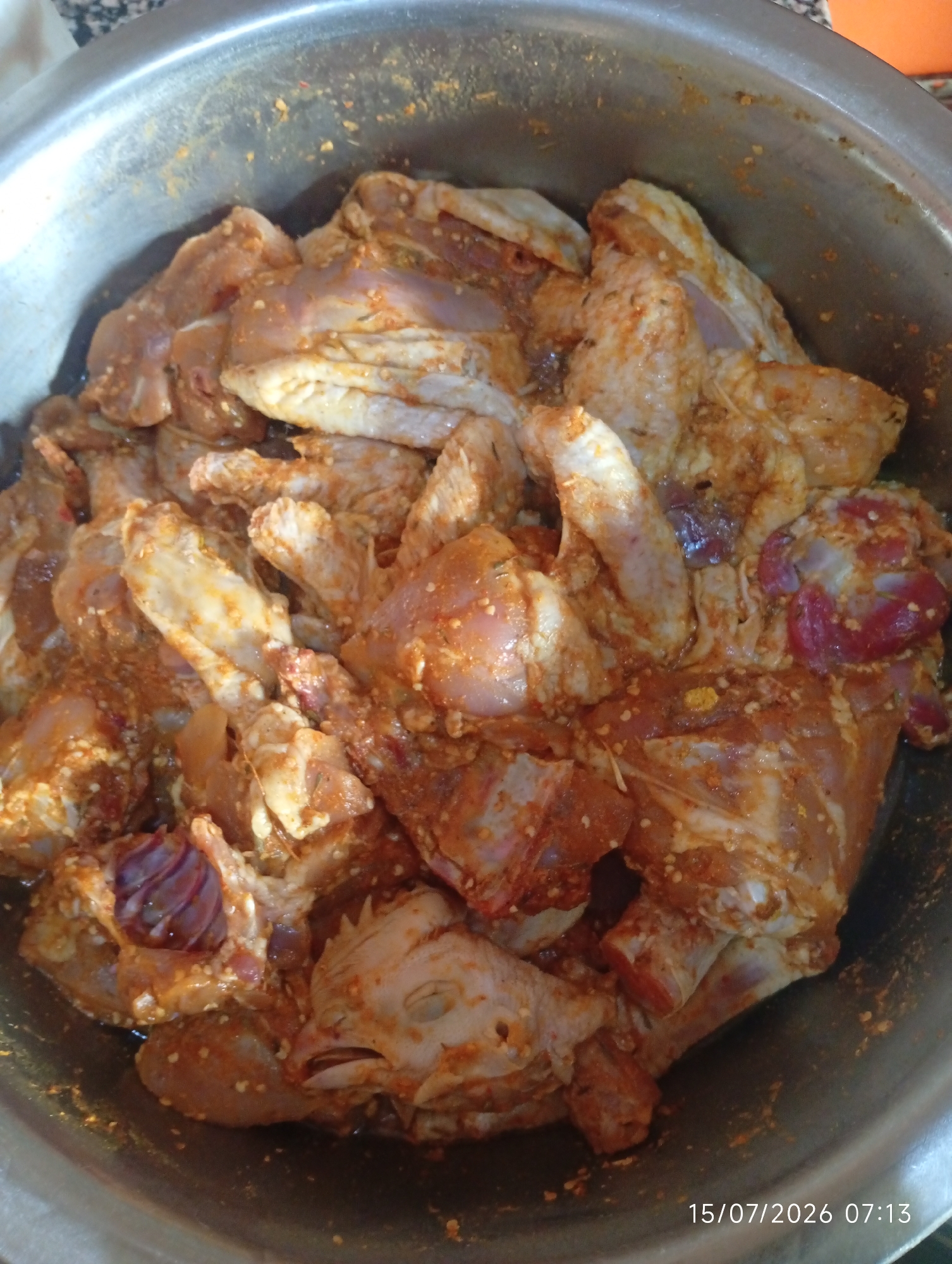 A close-up, overhead shot displays a stainless steel bowl filled with raw, cut chicken pieces that have been marinated in a reddish-orange spice mixture. The chicken pieces are of various sizes and cuts, including what appear to be wings, thighs, and potentially some giblets. The marinade coats the chicken unevenly, with some pieces showing more of the spice mixture than others. The bowl is large and metallic, with a slightly reflective surface. The background is blurred, but hints of a countertop with a patterned surface are visible. A timestamp in the lower right corner reads "15/07/2026 07:13". The image does not provide information about the specific location in Bana Stream, Nigeria, or any people.