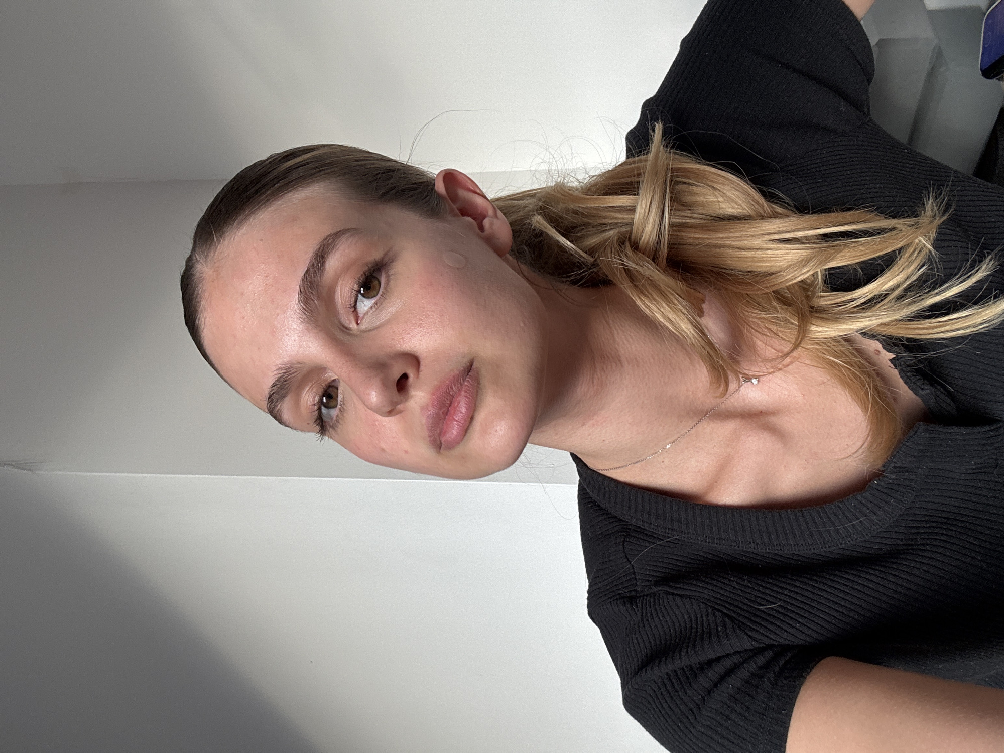 A young woman with light brown eyes and light blonde hair is depicted in a close-up, vertically oriented portrait. Her head is tilted to the right, and she looks towards the camera with a neutral expression. Her hair is styled in a way that pulls it back from her face, with a casual blonde ponytail or braid visible on her right side, and a few loose strands around her ear and neck.
A small, translucent, circular pimple patch is visible on her right temple/cheek. She wears a form-fitting, black ribbed-knit top with a rounded neckline. A delicate silver chain necklace with a small, star-shaped or diamond pendant rests on her collarbone. The background is a plain, light-colored (likely white or light gray) wall, suggesting an indoor setting. The lighting is bright and even. No specific activity is shown beyond posing for the photograph. There is no visible text in the image.