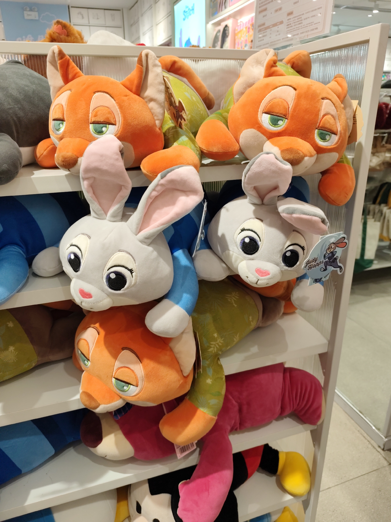 The image displays a retail shelf filled with stuffed animals, likely in a store. The merchandise includes plush toys of characters resembling Nick Wilde and Judy Hopps from Disney's Zootopia. Specifically, there are several orange fox plush toys with green eyes and pink inner ears stacked on top of each other. Interspersed among them are gray bunny plush toys with large black and white eyes and pink noses. One bunny toy has a tag attached that reads "Judy Hopps." The shelf also shows parts of other plush toys, including what appears to be a gray animal on the far left, and a pink plush leg extending from the bottom right. The background suggests a well-lit commercial setting with shelves and displays, typical of a toy store or department store. The retail environment is clean and organized. The image's metadata is not available to determine the exact location or time, but the presence of what appears to be Chinese signage in the background hints at a location in China. Without further information, pinpointing the exact location in Daqing, China, is not possible from the image alone.