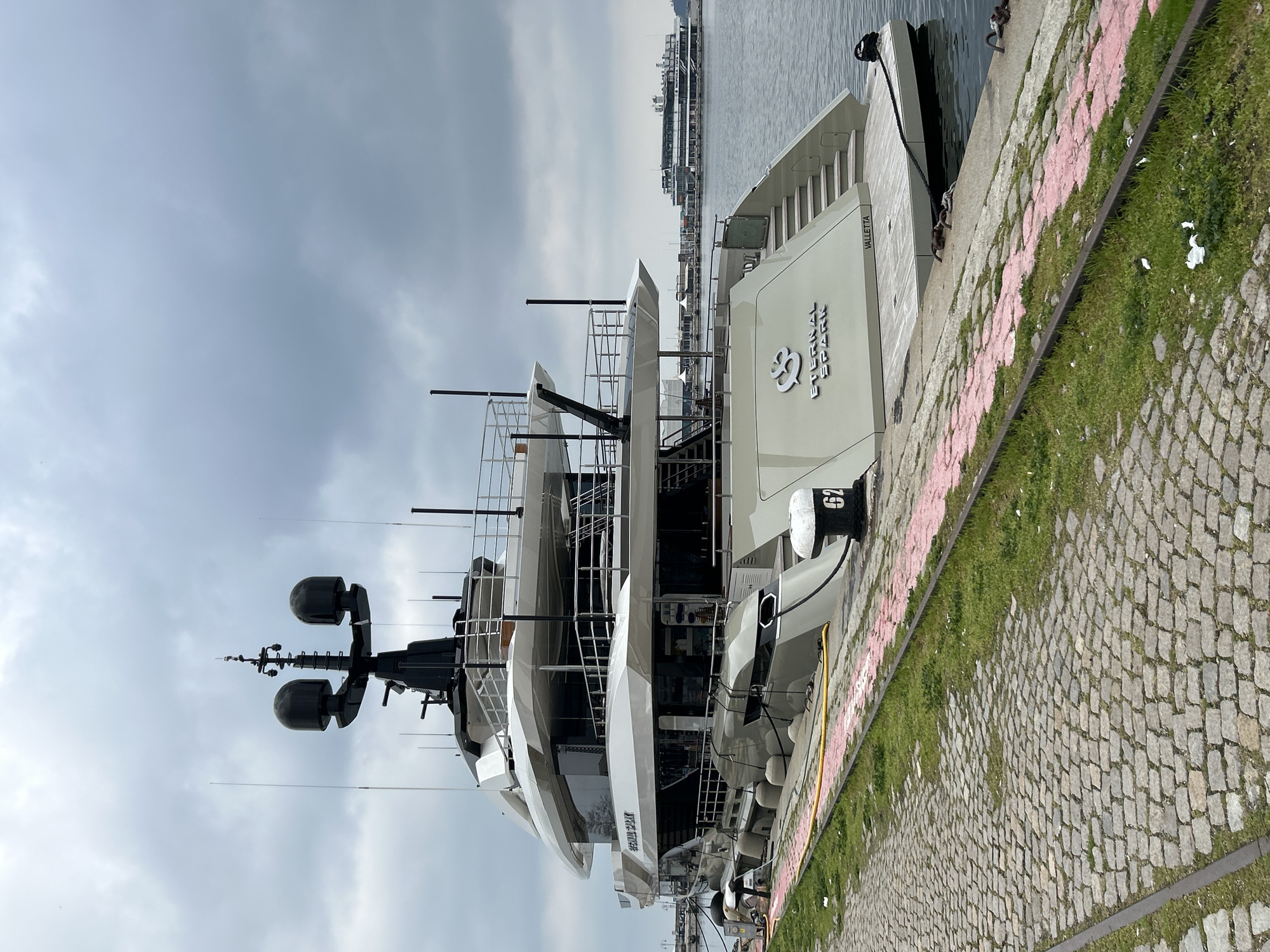 A medium, eye-level, outdoor shot captures the stern of a large yacht docked beside a stone and grass quay. The sky is overcast with grey clouds. The yacht, primarily white with black accents, is prominently featured, with the name "ESENCIA" displayed on its stern. Various deck levels and antennas are visible, adding to its size and complexity. The quay is made of cobblestones interspersed with grass and bordered by a low pink-painted concrete edge. In the distance, across the water, are buildings and possibly other docked vessels, suggesting an urban waterfront or harbor setting. The city is identified as VALLETTA, MALTA. The image is rotated 90 degrees counter-clockwise.