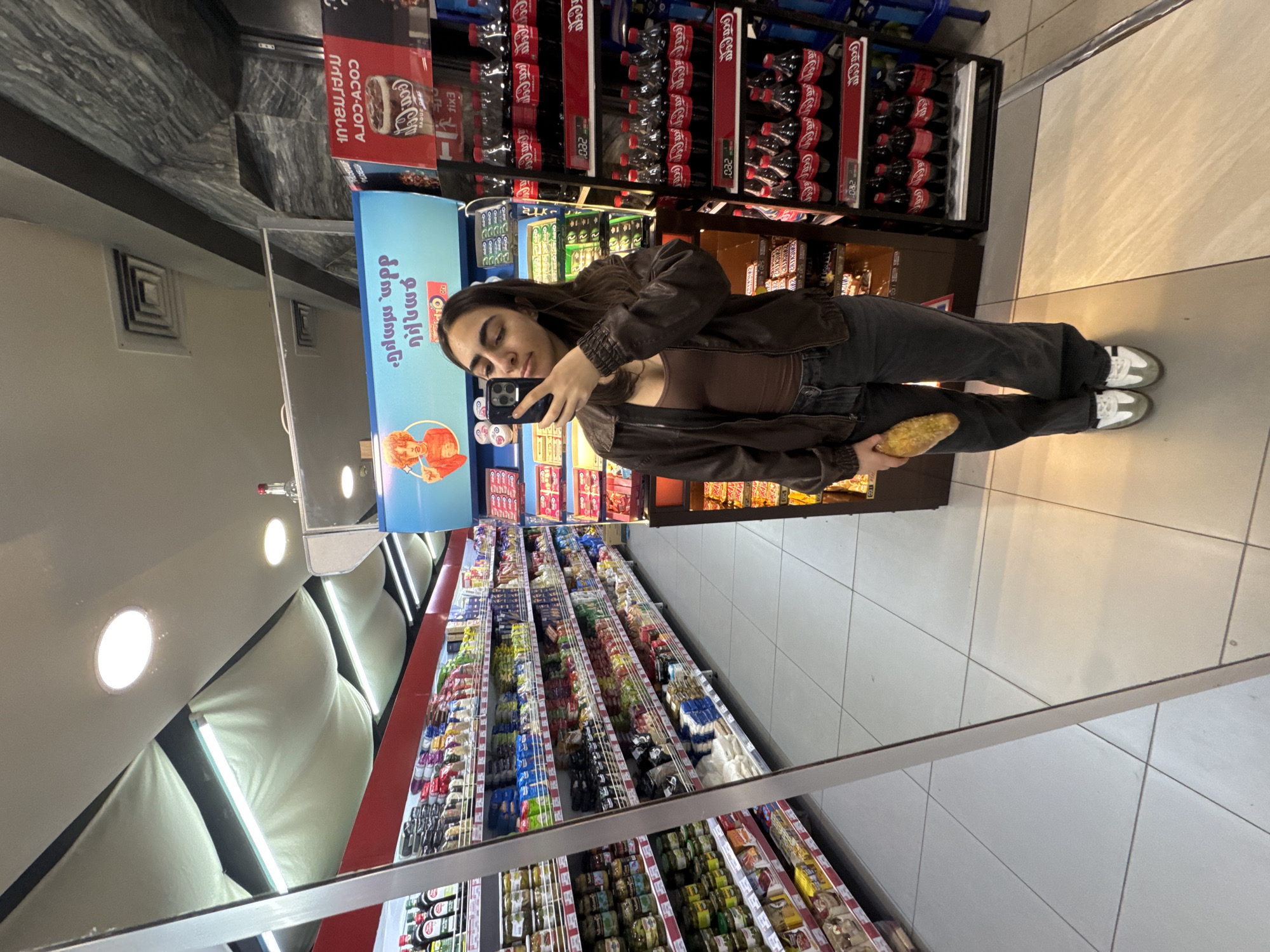 Here's a forensic description of the image:
**Scene:** A woman takes a photo of herself in a mirror inside a supermarket in Yerevan, Armenia. The reflection includes shelves stocked with various food products, beverages, and confectionery.
**Subject:**
* A woman with long dark hair, wearing a brown top and jacket with dark pants, and white-and-green sneakers. She is holding a phone in her right hand to take a selfie in a mirror. She also holds what appears to be a pastry in her left hand.
**Objects:**
* **Shelves:** Filled with various items, including Coca-Cola products.
* **Mirror:** Capturing the reflection of the woman and the surrounding environment.
* **Phone:** A smartphone is held by the woman, pointed towards her face, presumably to capture the selfie.
* **Pastry:** Held in the woman's left hand.
* **Ceiling lights**: Array of ceiling-mounted lighting fixtures
**Environment:**
* **Supermarket:** Evidenced by shelves stocked with a wide array of products.
* **Location:** Yerevan, Armenia; confirmed by the Armenian text displayed on some packaging.
* **Interior:** Aisle in the supermarket.