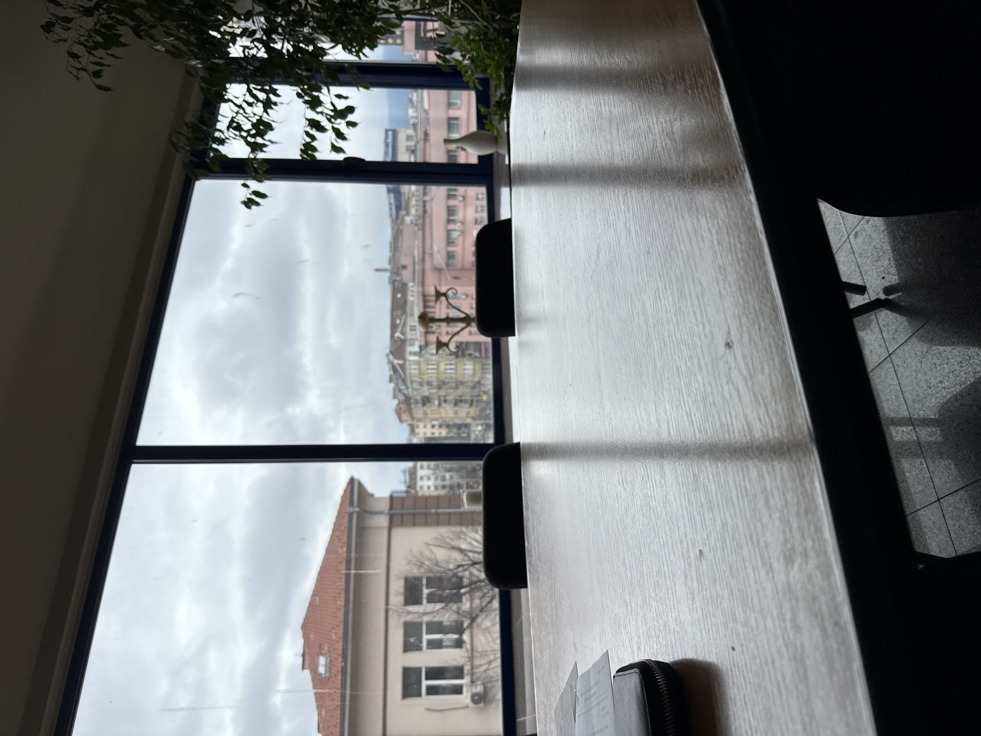 The image shows a view from a room in Sofia, Bulgaria. Outside the window is a cityscape with buildings, including a building with a red tiled roof and light-colored walls with visible windows and an air conditioning unit. Other buildings are visible in the distance. The sky is overcast. Inside the room, there is a long, light-colored table with at least two black chairs situated along the side facing the window. Some papers and a black wallet or small bag are visible on the table in the foreground. There is a leafy plant above the window. Tiled flooring is seen to the right of the table. The overall lighting suggests it is daytime.