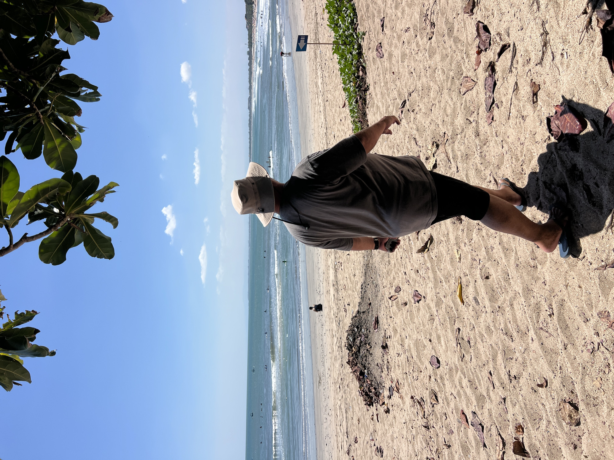 A man wearing a wide-brimmed hat, a gray t-shirt, and dark shorts walks along a sandy beach in Kuta, Indonesia. The man is seen from behind, with his right leg extended forward, and he is looking out towards the ocean. The beach is covered in sand, scattered with dried leaves and debris. In the distance, the turquoise ocean meets the horizon under a clear blue sky dotted with a few white clouds. Waves are breaking on the shore, and several surfers can be seen in the water. A small pile of debris is located on the sand to the left of the man. A blue sign with a white arrow pointing to the right is planted in the sand to the right of the man, with green foliage growing nearby. Trees with large green leaves hang over the top of the frame, casting shadows on the sand. The scene appears to be a typical sunny day on a popular tourist beach.