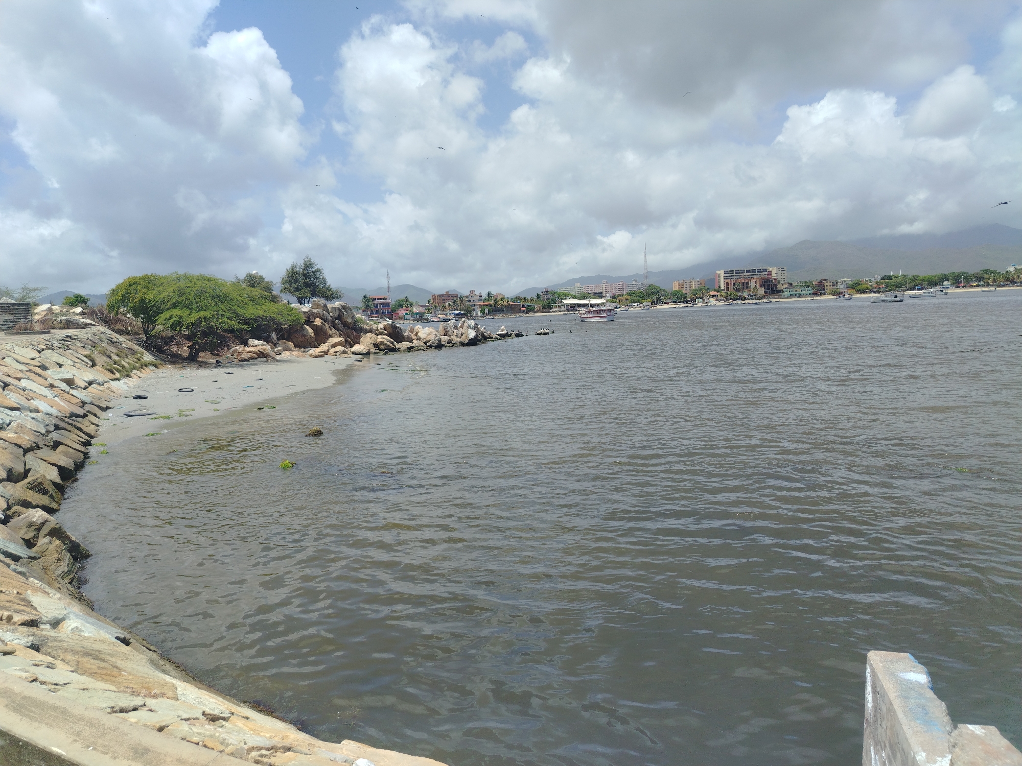 The image depicts a view of the coast in Juan Griego, Venezuela, under a partly cloudy sky. In the foreground, a rocky breakwater made of stacked stones lines the shore. Adjacent to the breakwater is a small sandy beach with some scattered debris, including a tire. The water in the bay is choppy and a murky dark gray. In the mid-ground, the coastline of Juan Griego is visible, featuring a collection of buildings, some of which appear to be hotels or residential structures, along with a few boats anchored in the water. A white pleasure boat with a red trim is moving in the bay. The background consists of rolling, green-covered mountains. Several birds are visible in the sky, and one appears to be in flight in the upper right corner of the image. A concrete structure is visible in the lower right corner of the frame.