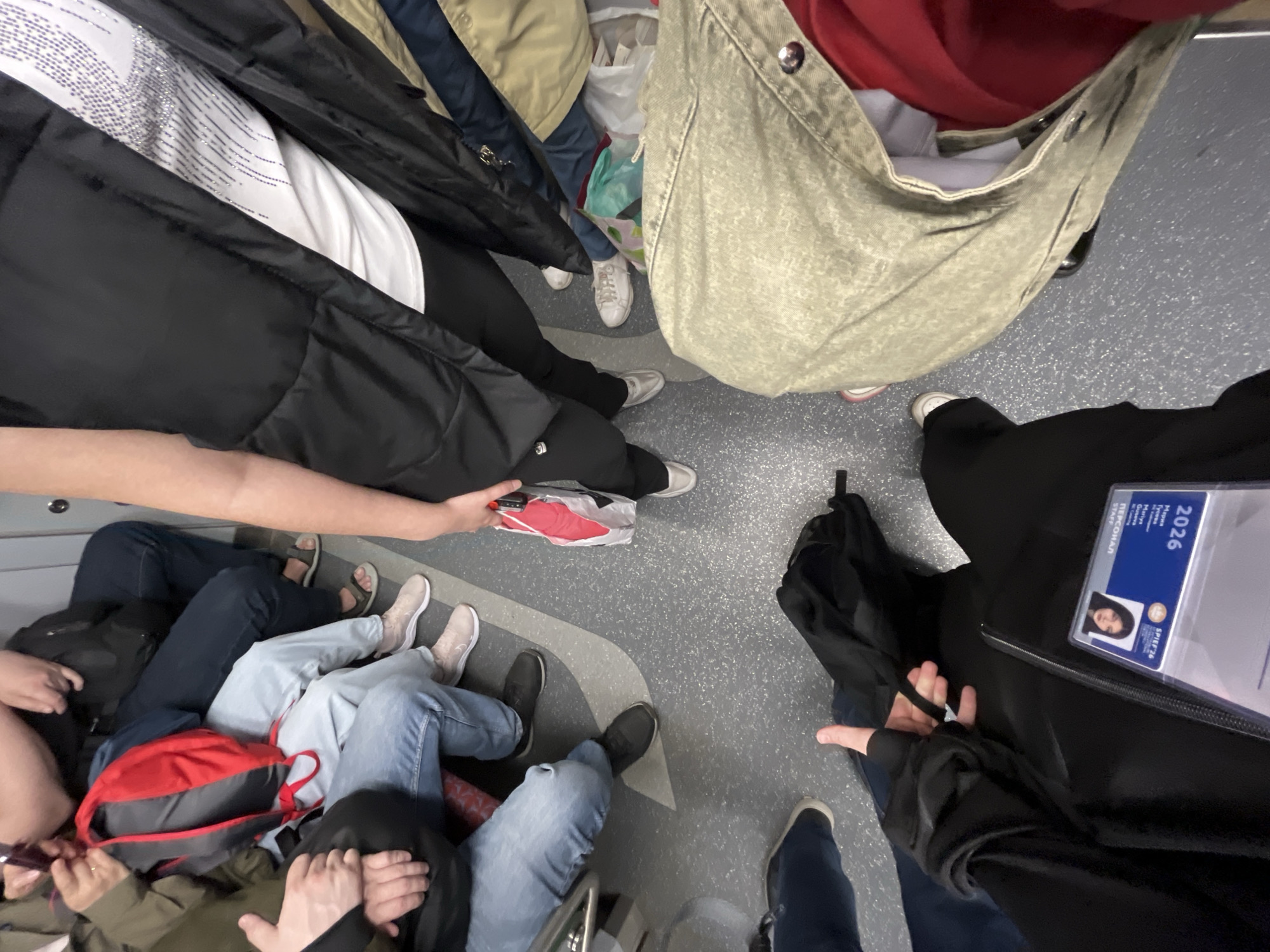 This image presents a high-angle view of a crowded public transport interior, likely a metro or bus, in Saint Petersburg, Russia. The perspective looks down onto a cluster of people, primarily showing their torsos, legs, and feet, suggesting a busy commuting or travel scenario.
Multiple individuals are present, with at least eight to ten pairs of legs and various types of footwear visible, including white, beige, and black sneakers, and sandals. Clothing includes blue jeans, black trousers, and jackets in black, tan, and red. One person wears a white top with silver embellishments.
Various personal items are scattered or carried by the individuals. These include a large, light-green canvas tote bag, a red and grey backpack, a small black backpack, and a white plastic bag. One person is seen holding a red object, possibly a power bank, within a clear plastic bag. An umbrella is also visible, held by one of the individuals.
A notable detail is a blue identification card displayed in a clear plastic holder. The card features a photograph of a person with dark hair and displays the text "2026", "STAFF", and Cyrillic script. The Cyrillic text reads "Персонал" (meaning "Personal" or "Staff") and appears to include the name "Ирина Галуш Мария" (Irina Galush Maria), consistent with the provided location context.
The floor of the vehicle is grey with speckled white markings, featuring a curved lighter grey line, indicative of an aisle or standing area. The surrounding panels are light grey. The image captures a moment of collective travel in a confined, bustling space.