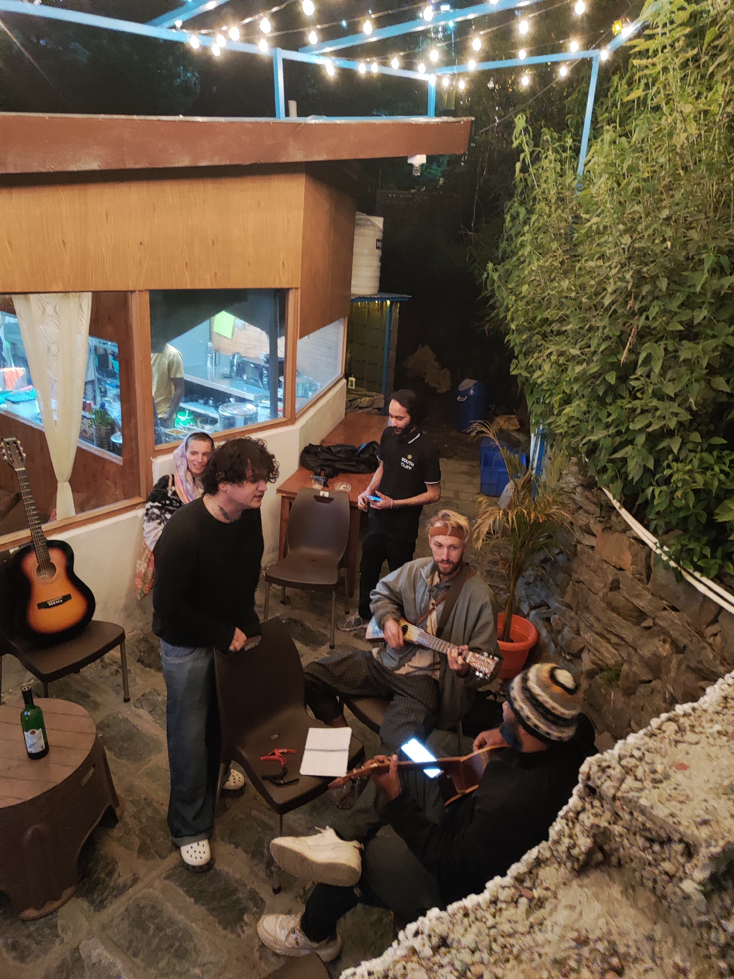 This media file captures an informal evening gathering in an outdoor setting in Haini, India, likely a patio or courtyard. The scene is illuminated by numerous string lights strung across an overhead blue metal frame, indicating it is nighttime.
Several individuals are present, with music being a central activity. In the foreground, two men are seated and playing acoustic guitars. One man, with blonde hair and a headband, is strumming a light-colored acoustic guitar. Next to him, another man, wearing a striped beanie and a black top, plays a darker acoustic guitar, with a lit phone screen visible near his instrument, possibly for lyrics or chords. Behind them, a man in a black polo shirt, featuring the text "SOUTH CLIFF," stands and looks at his phone.
To the left, near a wooden cabin-like structure with large windows, a man with curly dark hair and a black top stands, observing the musicians. Beside him, a woman with a head covering and a patterned shawl is seen smiling towards the group. Through the window of the wooden structure, another person in a yellow top is partially visible inside.
The environment features stone flooring and a textured stone wall adorned with lush green foliage on the right side. A potted plant is placed near the seated musicians. Furniture includes several brown plastic chairs and a small round table holding a green bottle. Another acoustic guitar rests on a chair near the wooden structure. Blue plastic bins are visible in the background. The overall mood appears relaxed and social, suggesting an impromptu jam session or a casual musical performance among friends.