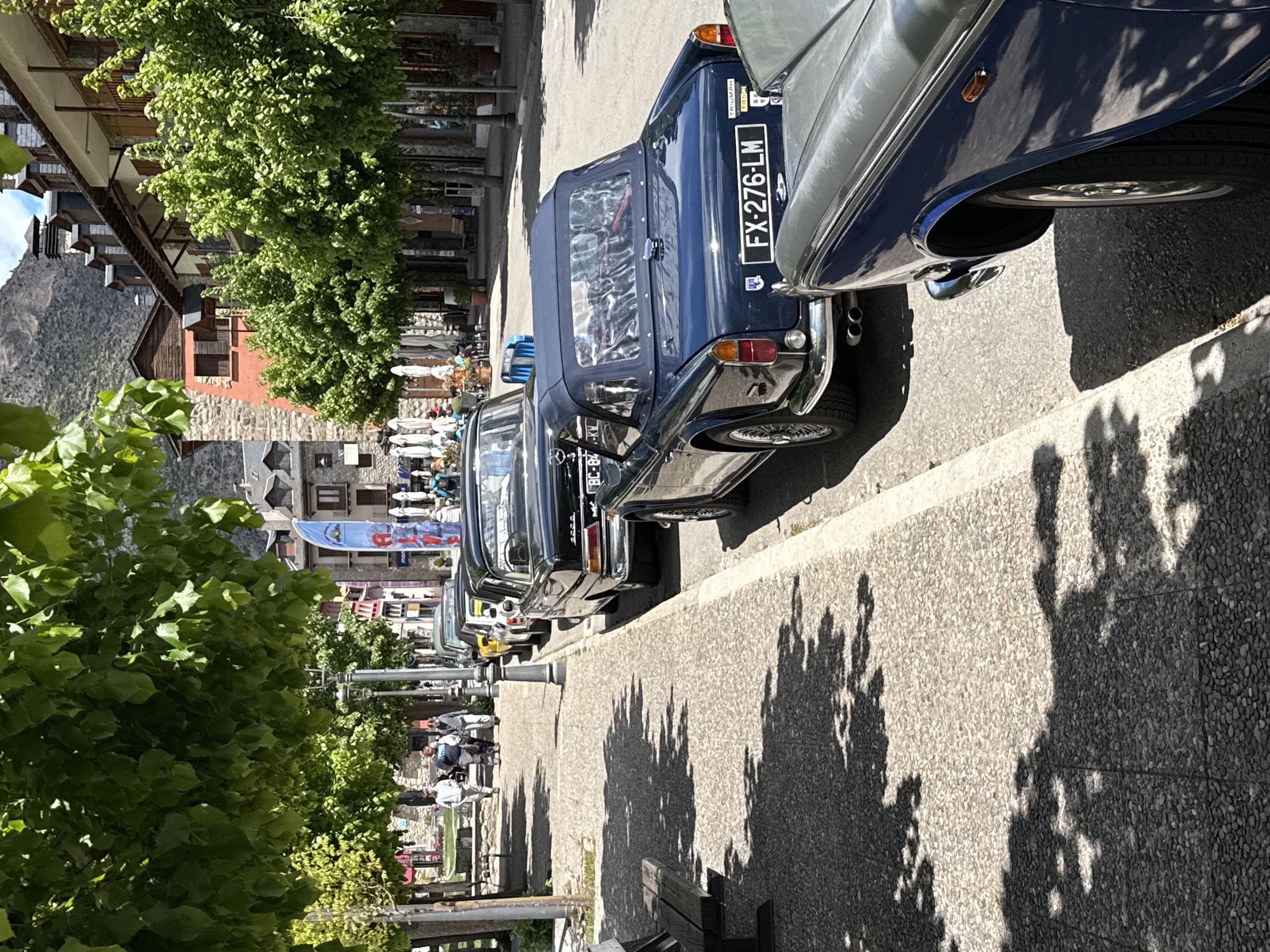 A row of vintage cars lines a cobblestone street in Benasque, Spain, casting long shadows from the surrounding trees. The blue Triumph, with license plate FX-276-LM, is partially obscured by a silver car. Along the street, pedestrians stroll past shops and cafes under the shade of trees. Buildings with traditional stone and wooden architecture line the street, set against a backdrop of distant mountains.