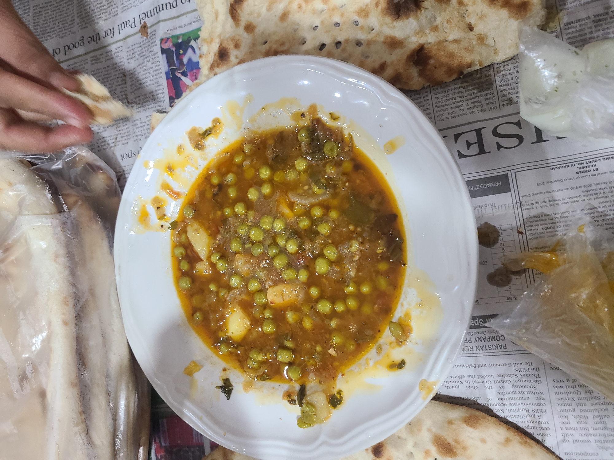 The image shows a plate of matar aloo (peas and potatoes curry) surrounded by naan bread and yogurt. The dish is served on a newspaper, possibly in a restaurant or home setting. A hand is seen reaching for a piece of naan bread.