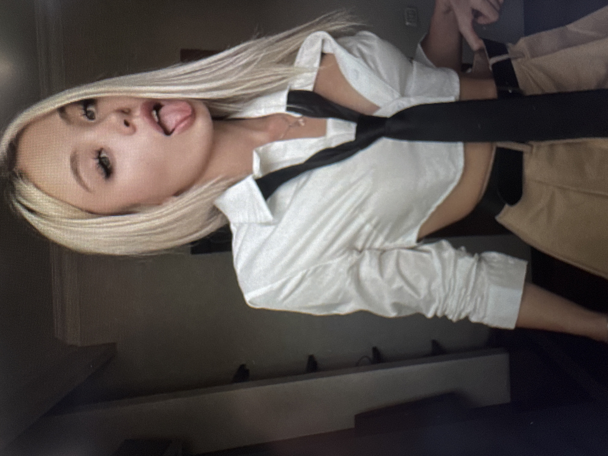 A young woman with long, straight blonde hair is featured in an indoor, chest-up shot. Her head is tilted, and she gazes directly at the camera with a playful expression, sticking her tongue out. She has noticeable dark eyelashes and appears to be wearing makeup. Her attire consists of a white, collared, long-sleeved cropped shirt, which reveals a small portion of her midriff. A black tie is worn loosely around her neck, draped over her shirt. She is also wearing light brown or tan pants. In the upper right area of the image, a hand, possibly belonging to another person or her own, is partially visible near her waist, seemingly touching her trousers. The background is a dark, indistinct wall with a lighter ledge or shelf running horizontally below it. The lighting is consistent with an indoor setting, likely artificial. The overall mood conveyed is one of confidence and lightheartedness. No text is visible in the image.