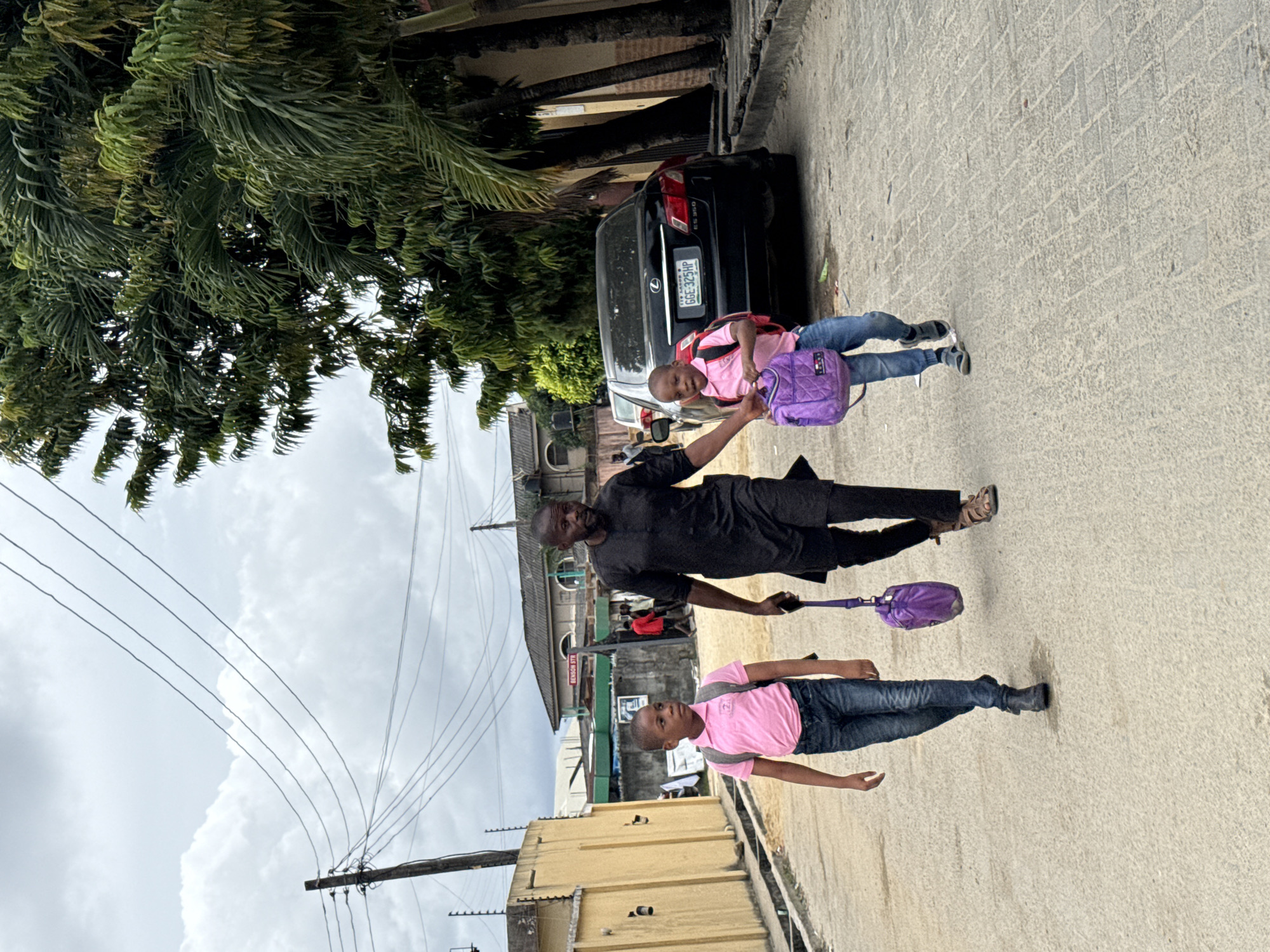 The image shows a man and two children walking on a paved street in Ado, Nigeria. The man is wearing a black outfit and sandals. He is holding a purple bag. One child is wearing a pink shirt, blue jeans, and black shoes and carrying a backpack. The second child wears a pink shirt and blue jeans and is being led by the man. A black car is parked on the side of the road. The background includes trees, buildings, and electrical wires. A sign reads "BENSON STR." The sky is overcast.