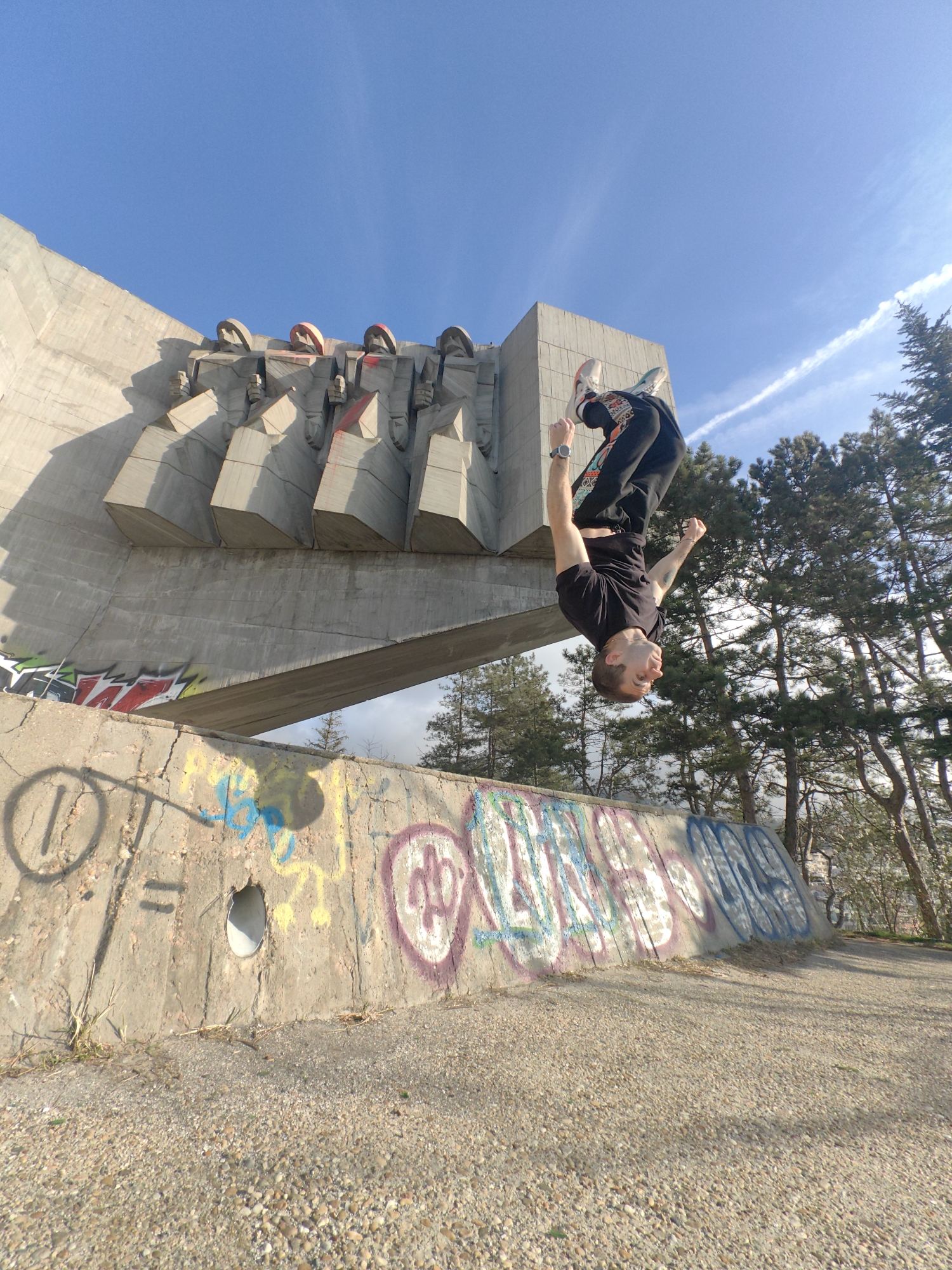 A male individual is captured mid-air performing a backflip against the backdrop of the Monument to the Bulgarian-Soviet Friendship in Varna, Bulgaria. The monument, a large brutalist concrete structure, occupies the upper-left of the frame, featuring stylized, block-like figures, some of which exhibit red spray paint. A lower concrete wall, heavily covered in multicolored graffiti, spans the foreground. The individual, dressed in a black t-shirt, patterned black trousers, and white athletic shoes, is inverted with arms raised. To the right, numerous tall evergreen trees rise towards a clear blue sky marked by white contrails. The ground is covered in light-colored gravel.