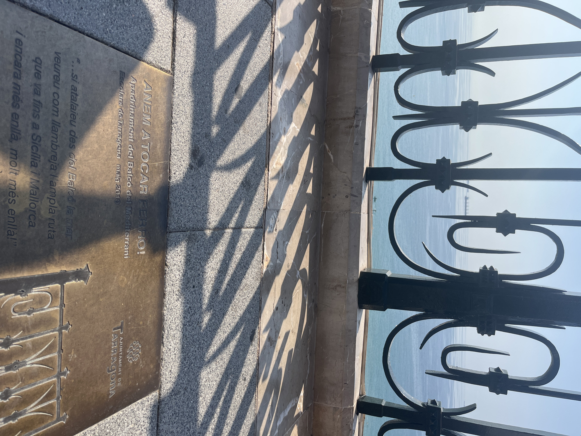 The image shows a bronze plaque embedded in the ground, likely a memorial or informational marker, with text in Catalan. The plaque is situated outdoors, on a paved surface, with a stone ledge and a wrought-iron railing visible behind it. Beyond the railing is a body of water, likely the Mediterranean Sea, with a ship on the horizon. Sunlight casts long shadows from the railing onto the pavement. The plaque itself has a title "ANEM A TOCAR FERRO!", followed by "Apadrinament del Balcó del Mediterrani" and "Ecolars de Tarragona maig 2010". Below this, there is a quote: "...si atalaieu des del Balcó la mar veureu com llambrequer l'ampla ruta que va fins a Sicília i Mallorca i encara més enllà, molt més enllà!". To the right of the text, there is a logo for "AJUNTAMENT DE TARRAGONA", the City Council of Tarragona. The overall scene suggests a viewpoint or promenade in Tarragona, Spain, offering a view of the sea.