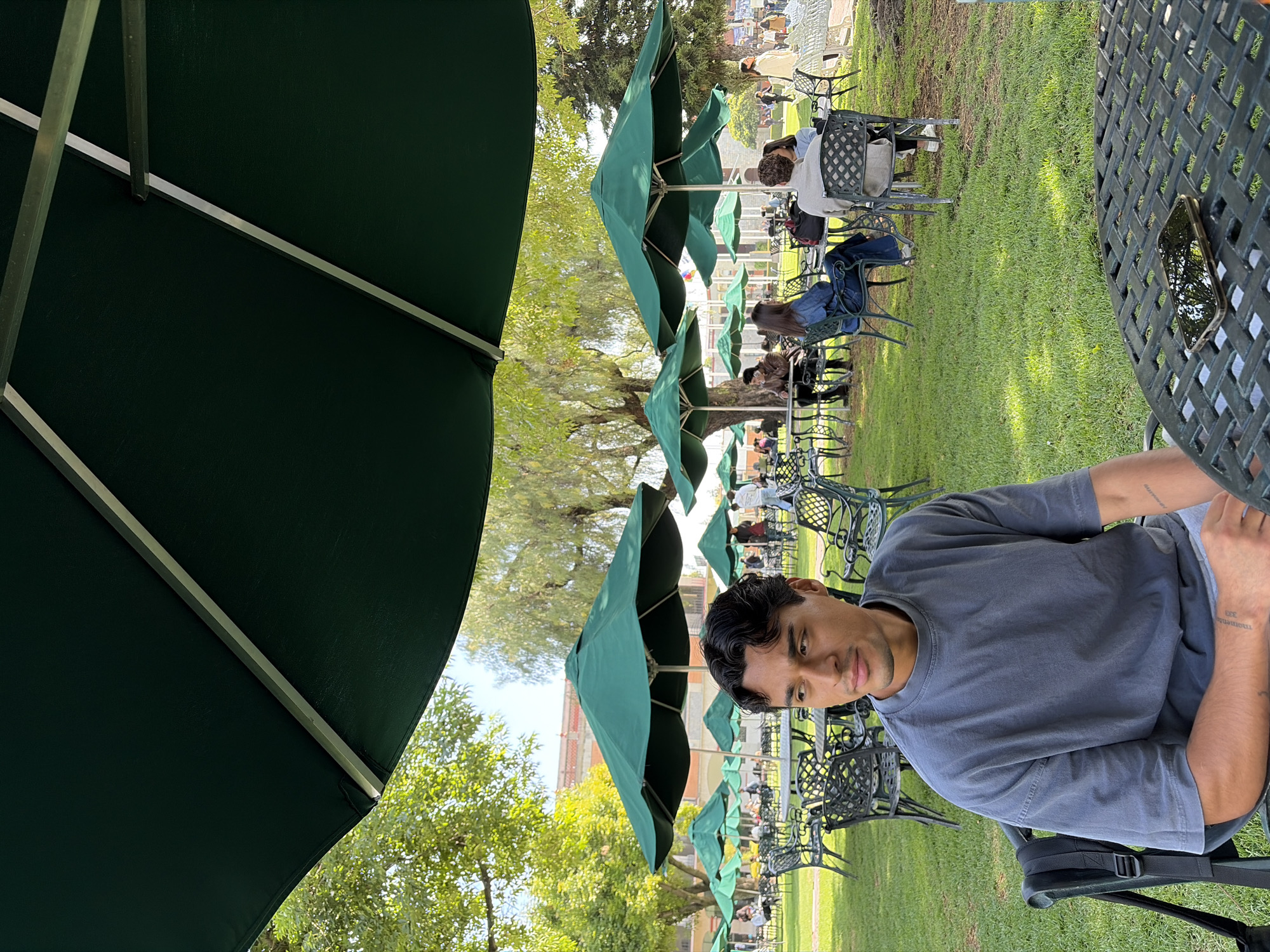 The image shows a man sitting at an outdoor table under a large green umbrella. He is wearing a grey t-shirt. In the background, there is a row of identical green umbrellas covering a seating area on a grassy lawn. People are seated at various tables. The scene appears to be located in San Andrés Cholula, Mexico, based on the architecture of the buildings in the distance. A phone sits on the table next to him.