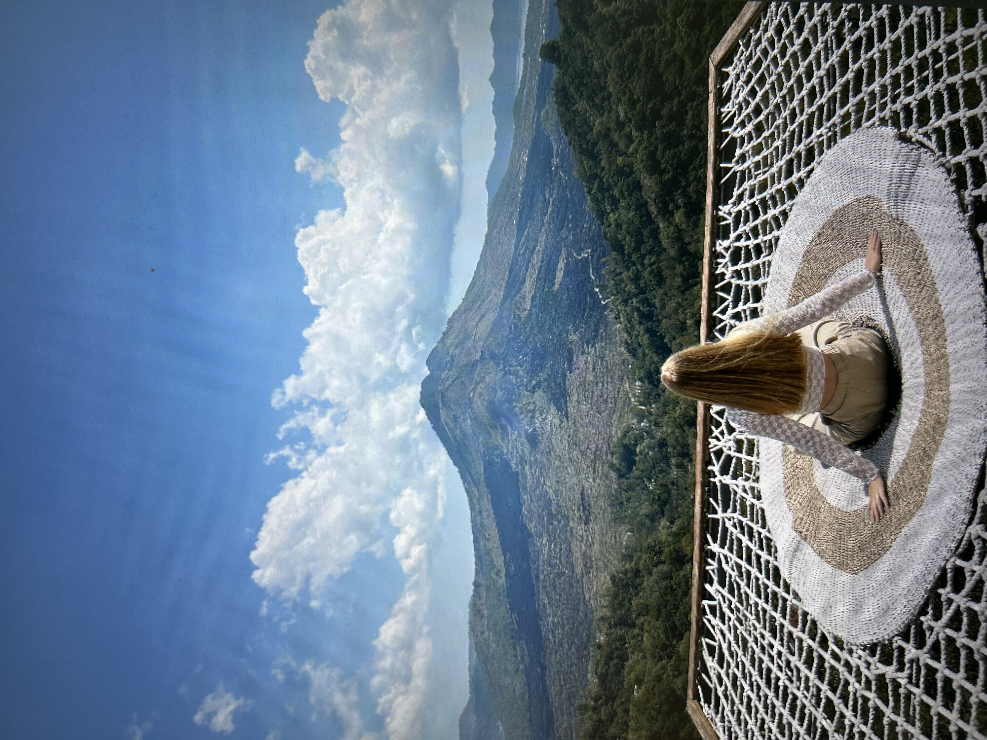 A woman with long, blond hair lies on her stomach on a circular striped pad placed within a rope hammock suspended in front of a mountain landscape under a partly cloudy sky. The sky is a pale blue with white clouds. The mountain is covered in vegetation and has a steep, rocky face. The woman wears a light-colored long-sleeved shirt and beige pants. She is reaching her hands out to the sides. The hammock is attached to a wooden frame. The photo appears to be taken at a high altitude, overlooking a vast landscape. This area can be found near Kuta Selatan, Indonesia.