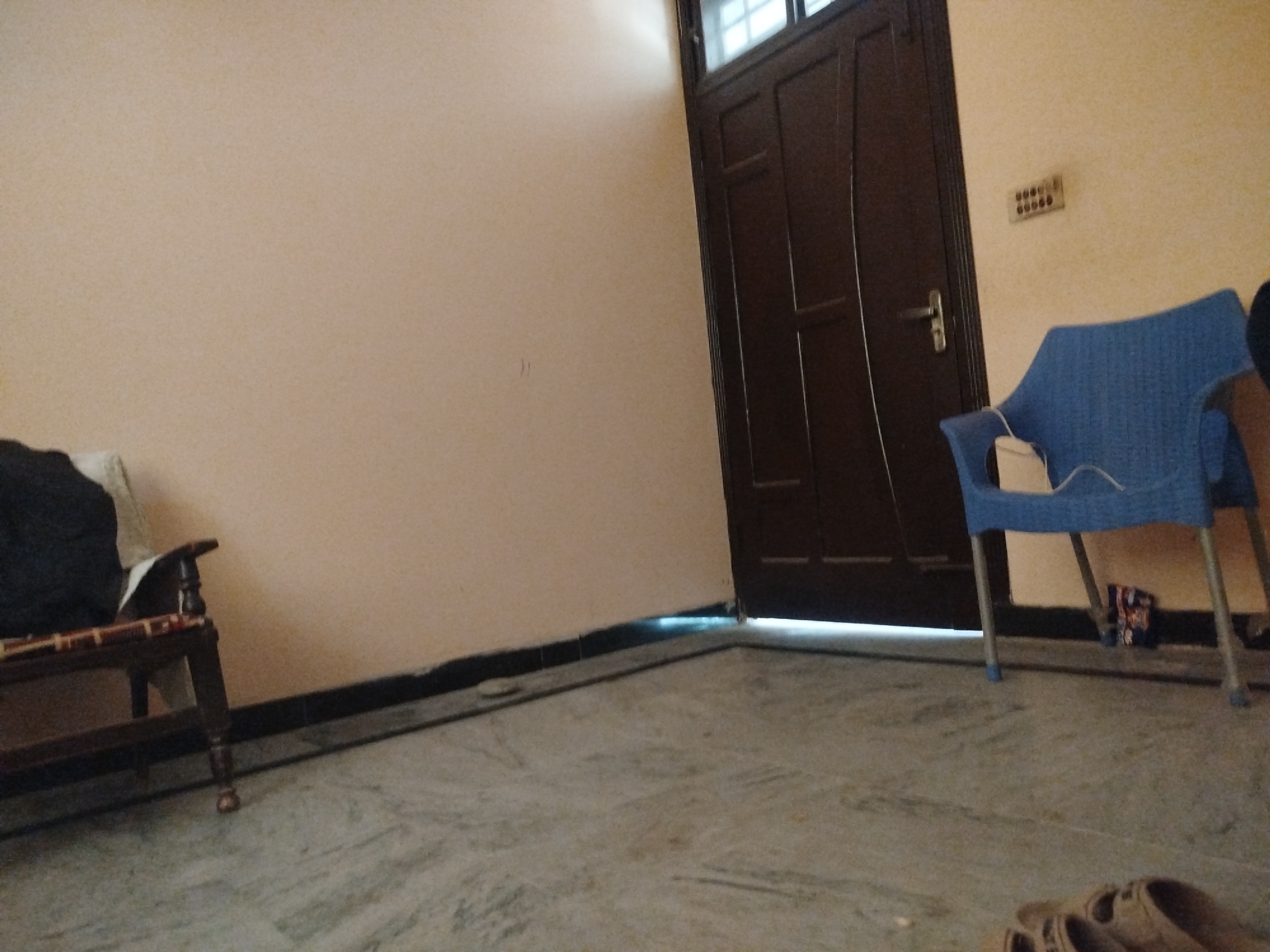 The image depicts an indoor scene, likely a room in a house in Lassan, Pakistan. The floor is made of marble or stone with a light grey and white marbled pattern. A dark brown wooden door with paneling and a silver doorknob is visible on the right side of the image, partially open, allowing light to enter from an unseen area. To the right of the door, a light yellow wall features an electrical outlet with multiple circular sockets. In the foreground, a blue plastic chair with silver legs stands on the floor. A small bag or packet is visible on the floor near the chair. On the left side of the image, a dark wooden bench with carved legs supports a cushion and what appears to be a dark fabric. The walls are painted a neutral, light beige color. The overall impression is that of a simple, functional living space.