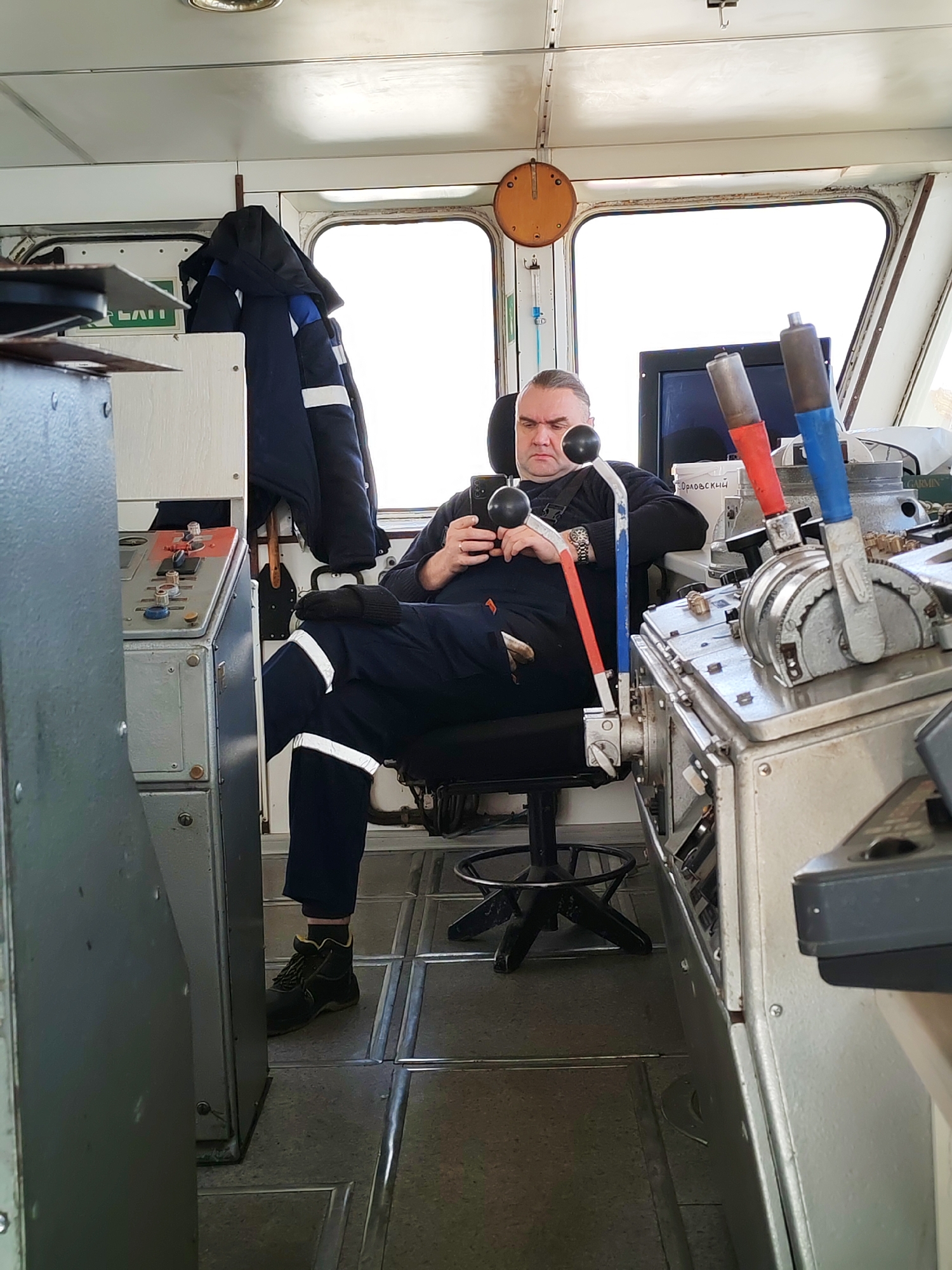 A man is seated in a swivel chair, engrossed in his phone. He's wearing a navy blue jumpsuit with reflective stripes, a black undershirt, and black boots. The scene appears to be inside the control room of a vessel, identifiable by the various control panels, levers with red and blue handles, a window with white frame, and a clock on the wall. The location is Unknown, Unknown.