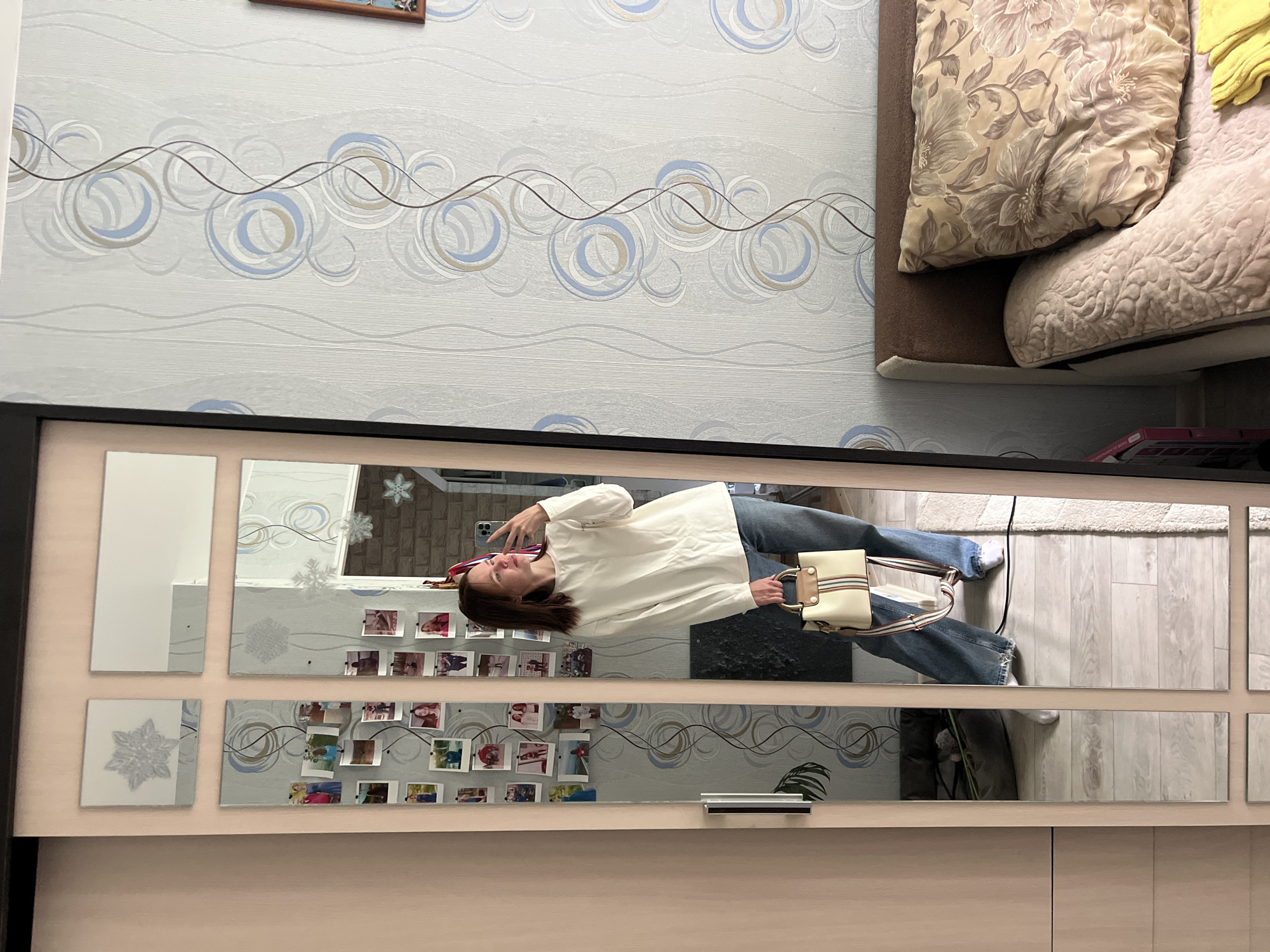 The image shows a woman standing in front of a wardrobe with a mirrored front, taking a picture of herself in the mirror with her phone. The woman is wearing a white blouse and blue jeans, and she is carrying a light-colored purse.
The wardrobe has multiple small square mirrors arranged in a grid pattern. Some mirrors display photos attached to them. The wall behind the wardrobe is decorated with a pattern of circular designs and horizontal lines. The top corner of a bed with patterned pillows is visible in the upper right corner of the image.
The location is Chaikovsky, Russia, as indicated by metadata.