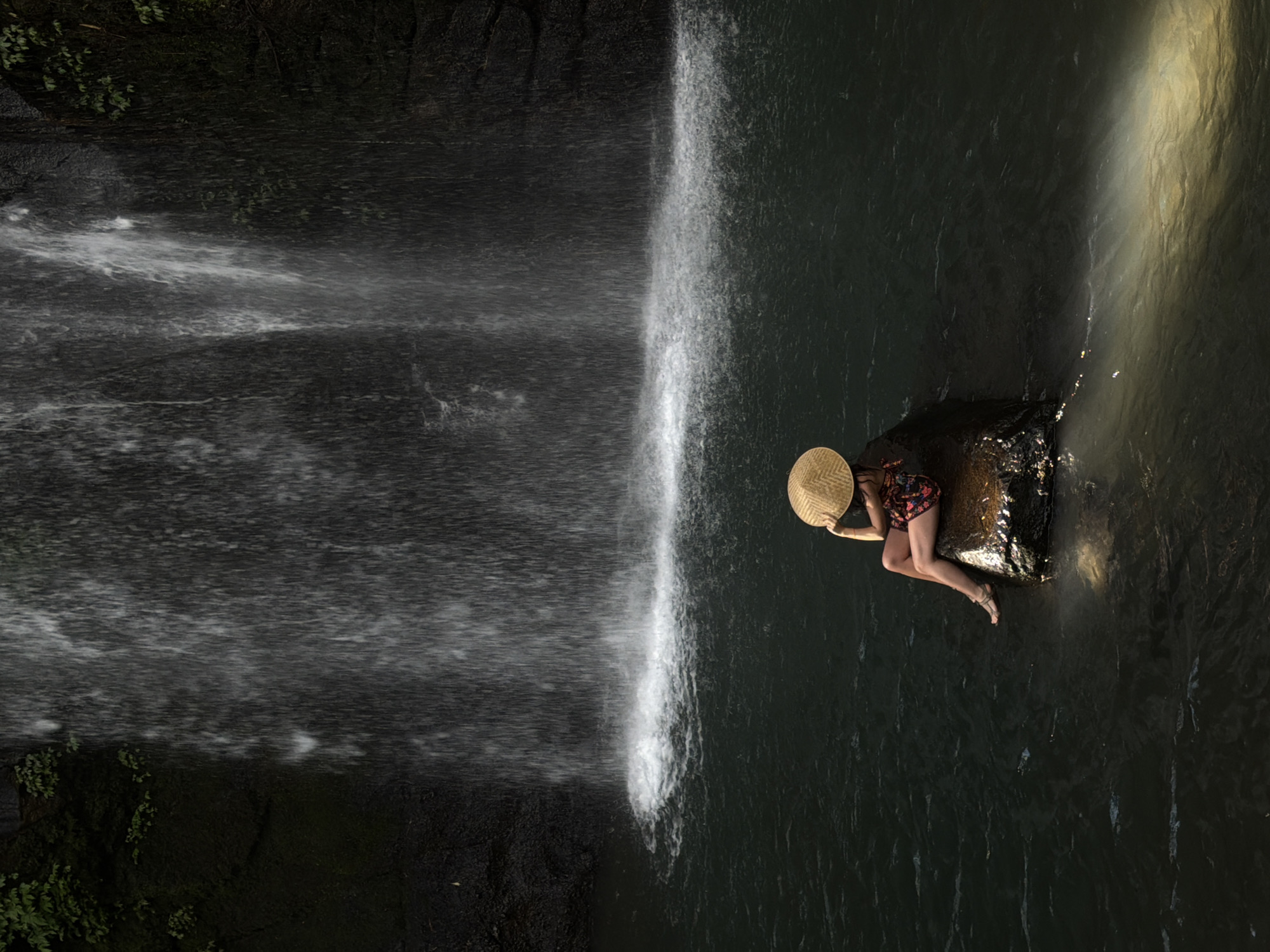 A woman wearing a wide-brimmed straw hat and a patterned dress sits on a dark, wet rock in the shallow water at the base of a waterfall. The waterfall cascades down a dark, mossy cliff face, with streaks of white water creating a misty curtain. The water at the base is dark and calm, with a bright patch of sunlight reflecting on the surface to the right of the rock. The woman is positioned with her back to the camera, looking towards the waterfall. Her left leg is bent and submerged in the water, while her right leg is extended. The scene appears to be a natural setting, possibly in Tegallalang, Indonesia, known for its rice terraces and waterfalls.