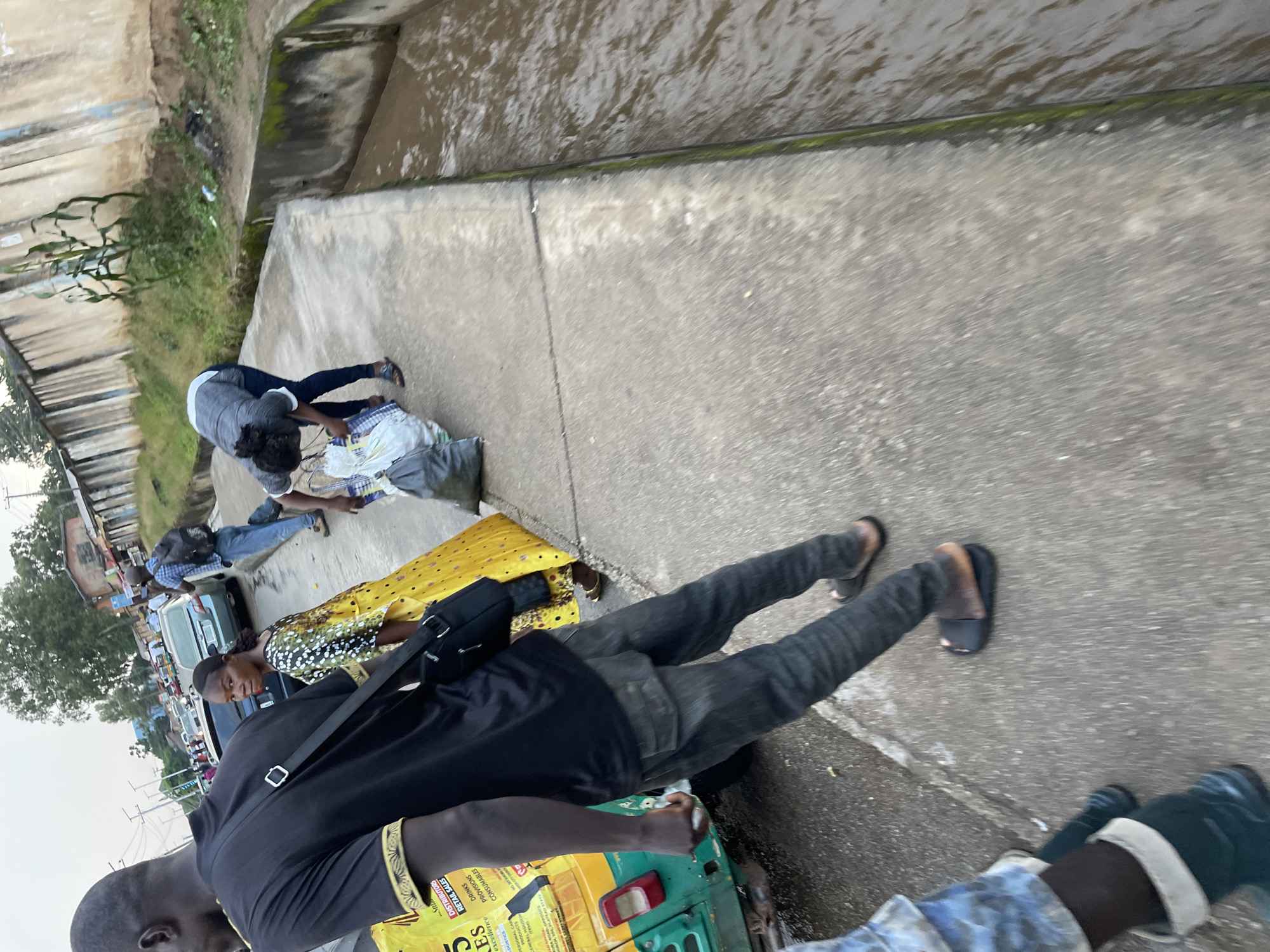 In Jos, Nigeria, a group of people are walking along a paved path next to a water channel. In the foreground, a man in a black t-shirt and grey jeans is walking away from the camera, carrying a black bag. To his left, a woman in a yellow and green patterned dress is looking back. Further down the path, another person is bent over, collecting bags. In the background, a few vehicles are visible on a street lined with buildings and vegetation. The sky is overcast.