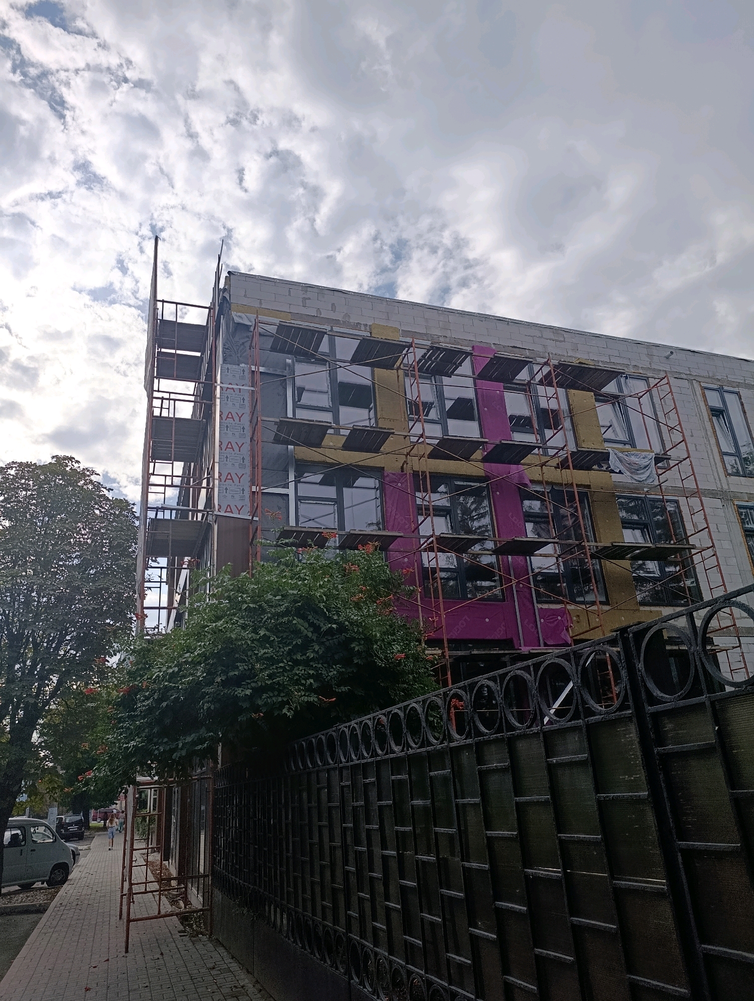 The image shows a construction site in the city of Cherkasy, Ukraine, with a building under renovation. The building, a multi-story structure, is partially covered with scaffolding and insulation materials. The facade features a combination of white brickwork, yellow insulation panels, and sections with magenta-colored material. Large windows with dark frames are visible across the building. Trees and foliage surround the construction site, with a dark metal fence in the foreground. A sidewalk with paving stones runs alongside the building, and a car is parked on the street. A few people are walking on the sidewalk in the distance. The sky is cloudy, with patches of blue visible.
