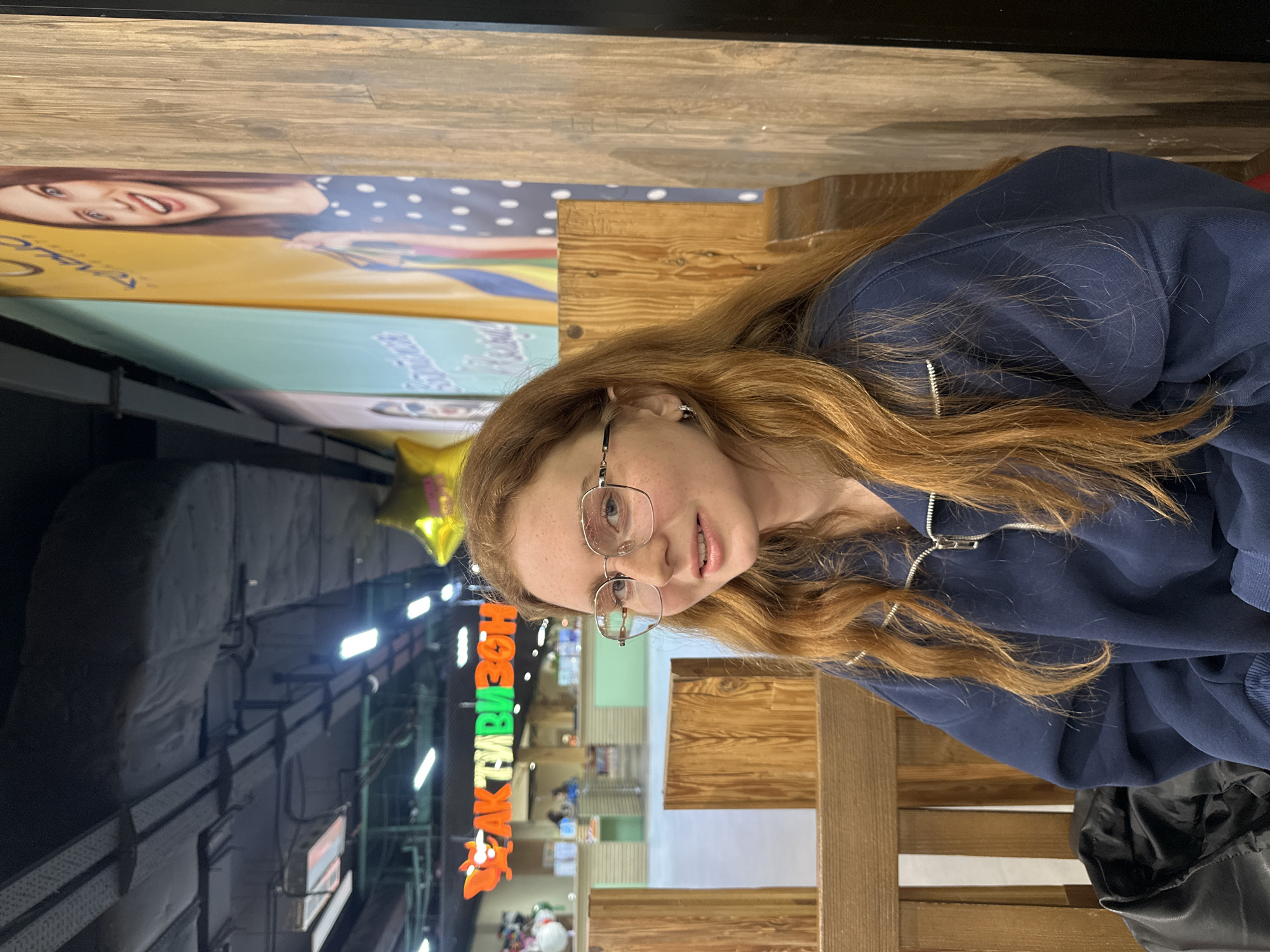 The image shows a young woman with red hair and glasses, wearing a navy blue zip-up jacket. She is seated at a wooden table in what appears to be a public space. In the background, there is a sign in Cyrillic script with brightly colored letters and orange cartoon characters. The sign reads "АКТИВИЗОН" (AKTIVIZON). Above the sign is a gold star-shaped balloon, along with overhead ventilation systems. The environment behind the sign seems to be a retail or entertainment area with multiple figures and displays. In the upper portion of the image there are commercial billboards and advertisement panels of women. Based on the “АКТИВИЗОН” sign, the location is in Kazan, Russia.