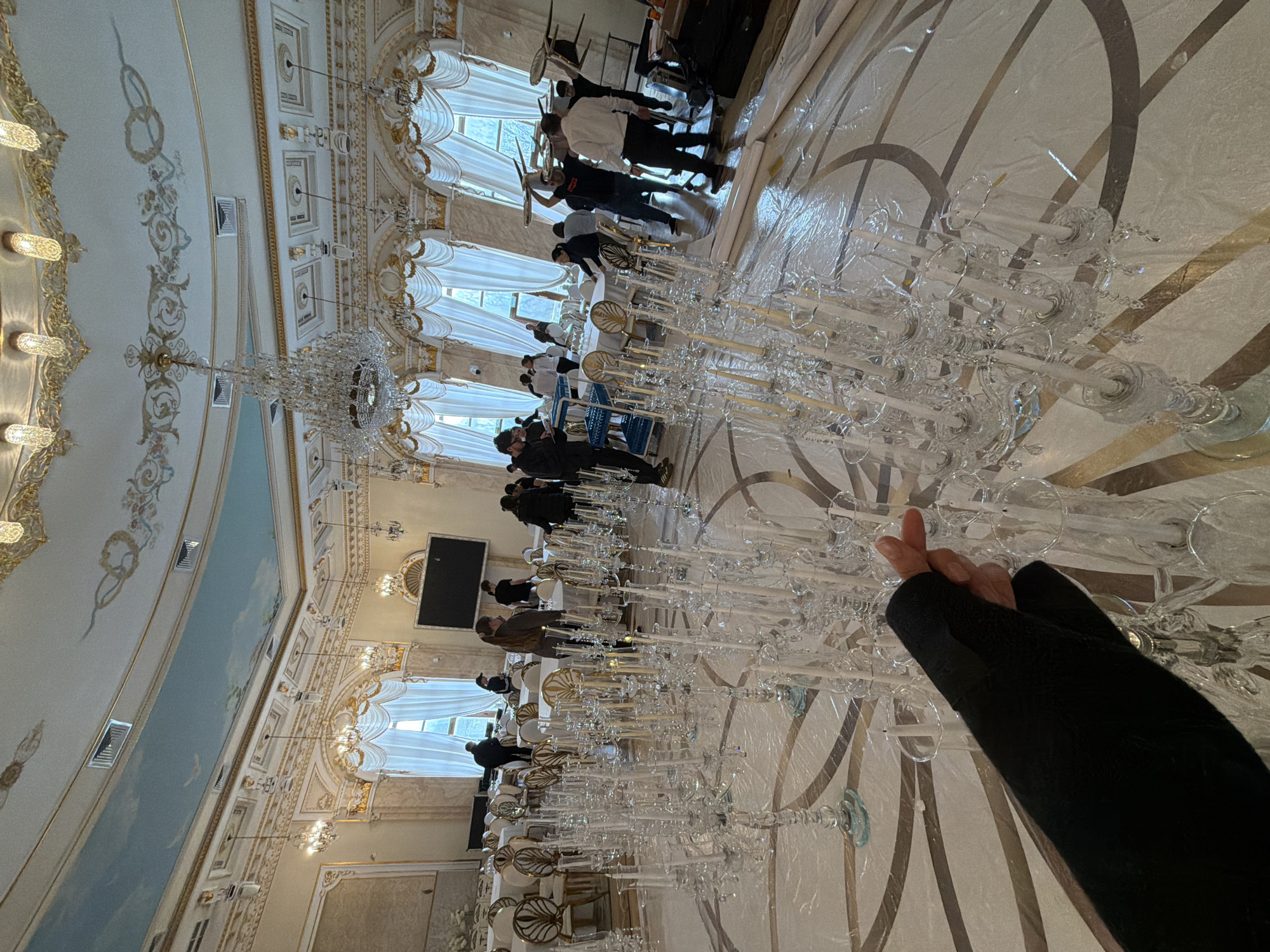 The image captures a scene inside a grand hall, likely within a historical building, possibly in Moscow, Russia, given the ornate architectural details reminiscent of Russian palaces. Several people are present, some appearing to be workers or staff, engaged in setting up or preparing the hall, while others are observing or supervising. The primary focus is on rows of tables, arranged for what seems like a formal event or banquet. These tables are covered with clear plastic sheeting and adorned with numerous tall, ornate candelabras. One person's hand in a black sleeve is raised near one of the candelabras. The architectural features include high ceilings with intricate detailing and a painted sky scene. Large windows draped with white curtains provide natural light. The hall also features a flat-screen television mounted on the wall. The floor is covered with a geometric pattern, partially obscured by the plastic covering the tables. The overall impression is that the hall is being prepared for a special occasion.