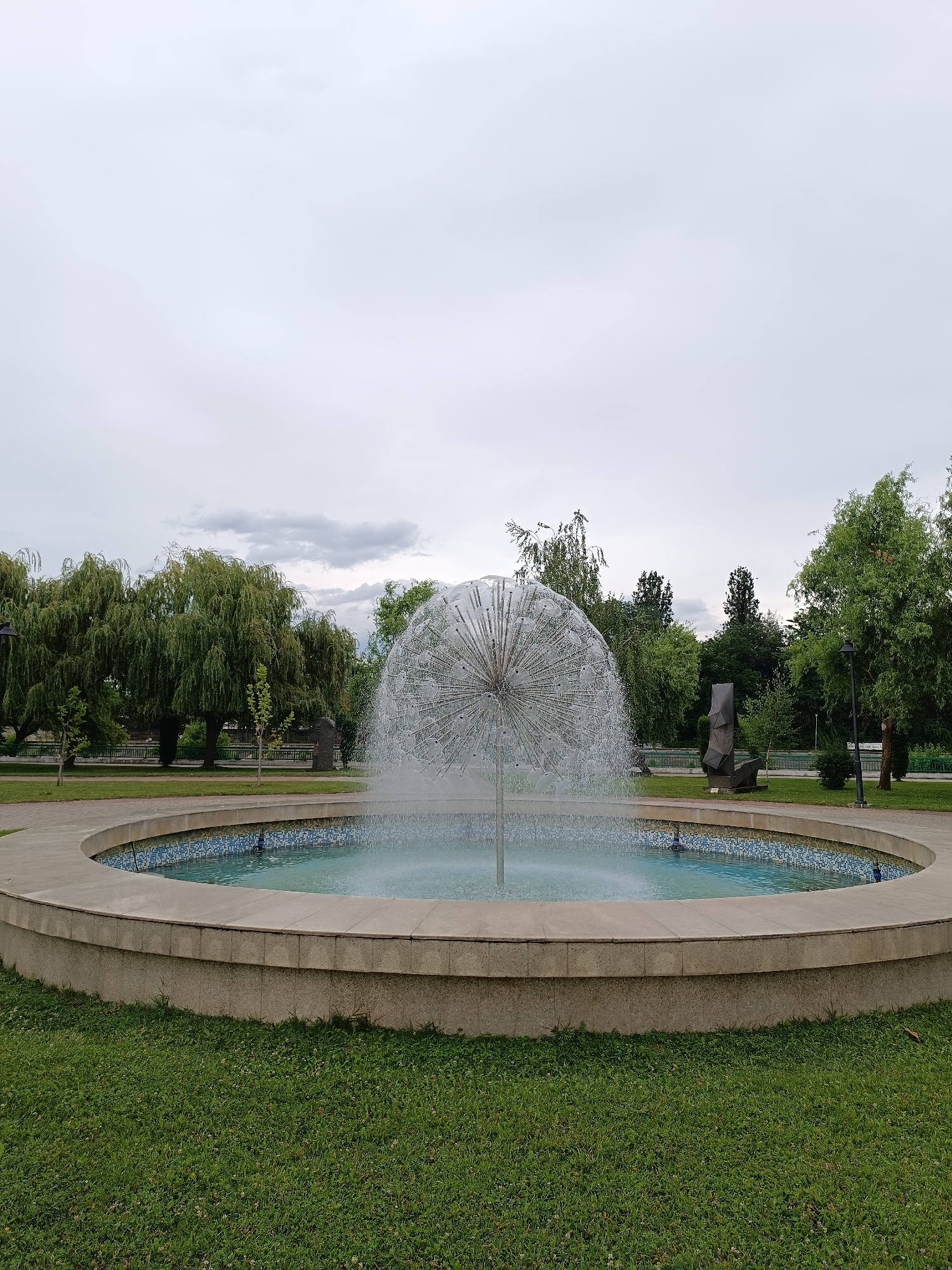 The image depicts a modern, spherical fountain spraying water in a circular pool. The pool is surrounded by a beige concrete rim, which is tiled with blue and white mosaic on the inside. Lush green grass covers the foreground, and a pathway curves around the fountain. In the background, several trees, including prominent weeping willows, line a park area. A contemporary sculpture, dark and abstract in form, stands to the right of the willows. The sky is overcast and grey. This fountain is located in the park near the Jiu River in Târgu Jiu, Romania.
