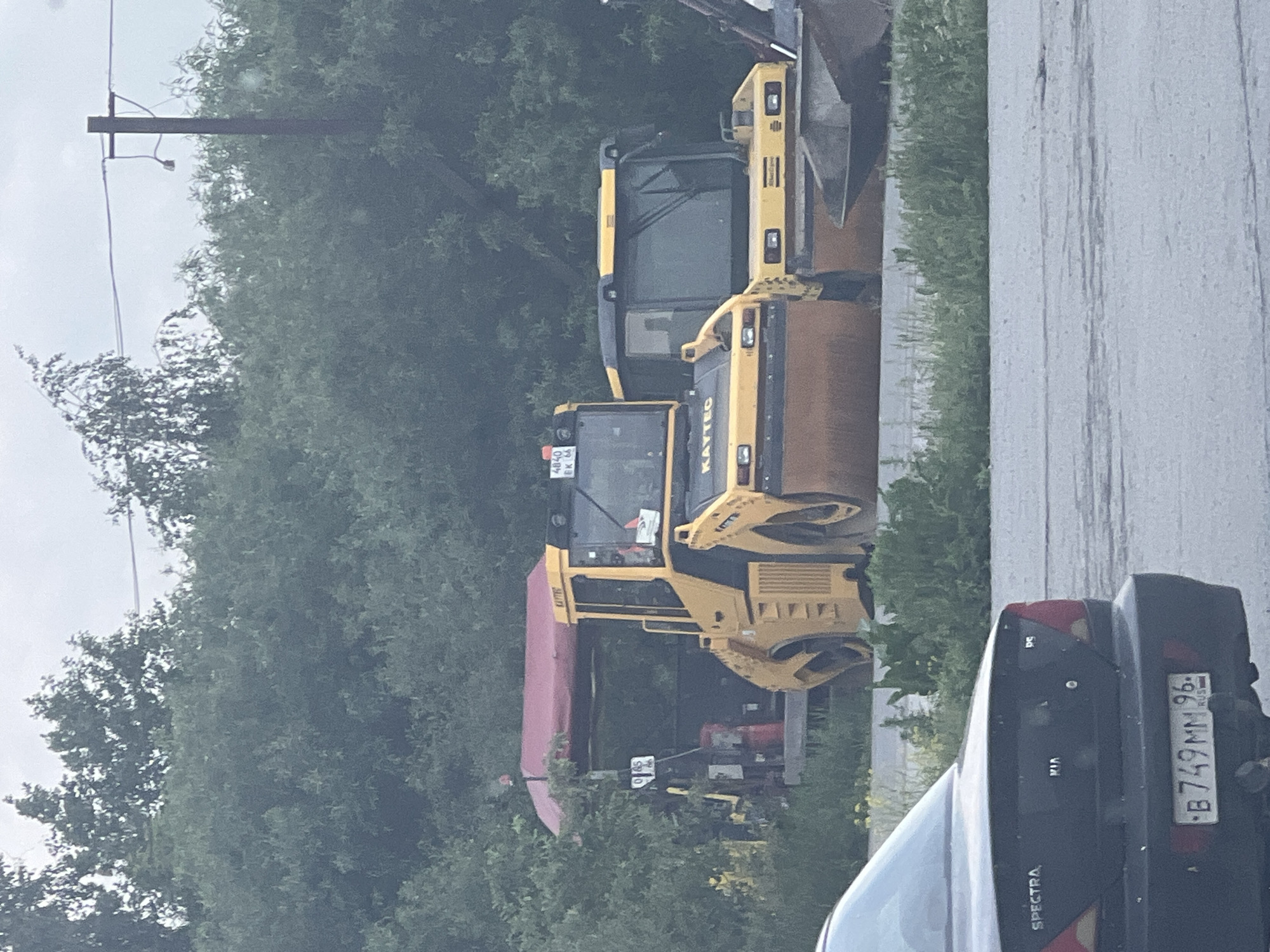 Two yellow road rollers are parked side-by-side in an outdoor lot, partially obscured by trees. The roller in the foreground is larger and features the brand name "KAYTEC" on its cabin. The roller behind it is slightly smaller and also has a "KAYTEC" logo. A red canopy structure is visible to the left of the rollers, with some equipment underneath. In the foreground, a dark-colored sedan is parked, with its rear end facing the viewer. The license plate of the sedan reads "B 749MM 96". The environment appears to be a somewhat overgrown area with green foliage and a cloudy sky.
